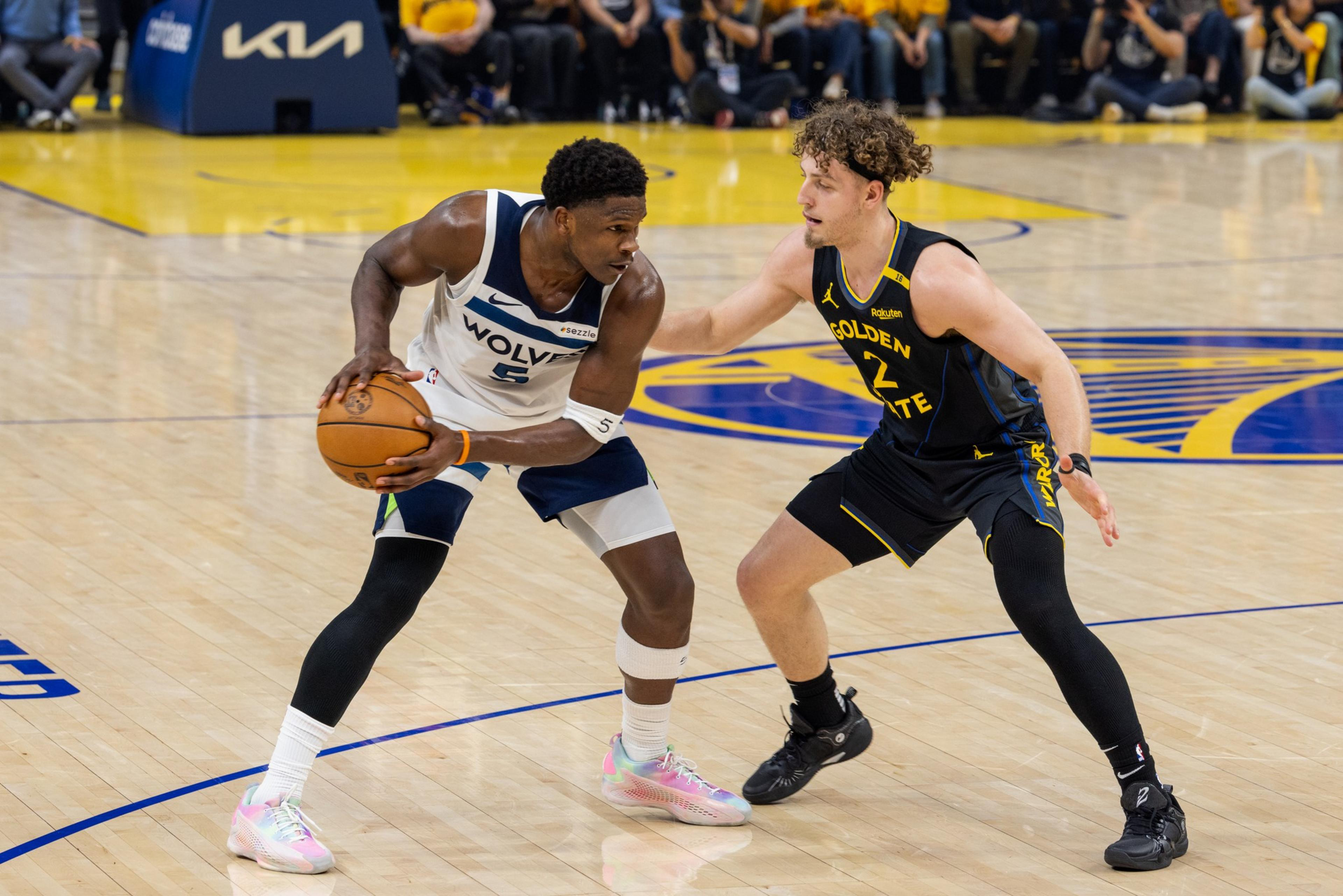 A basketball player in a Wolves jersey holds the ball, guarded closely by a player in a Golden State uniform on a court.