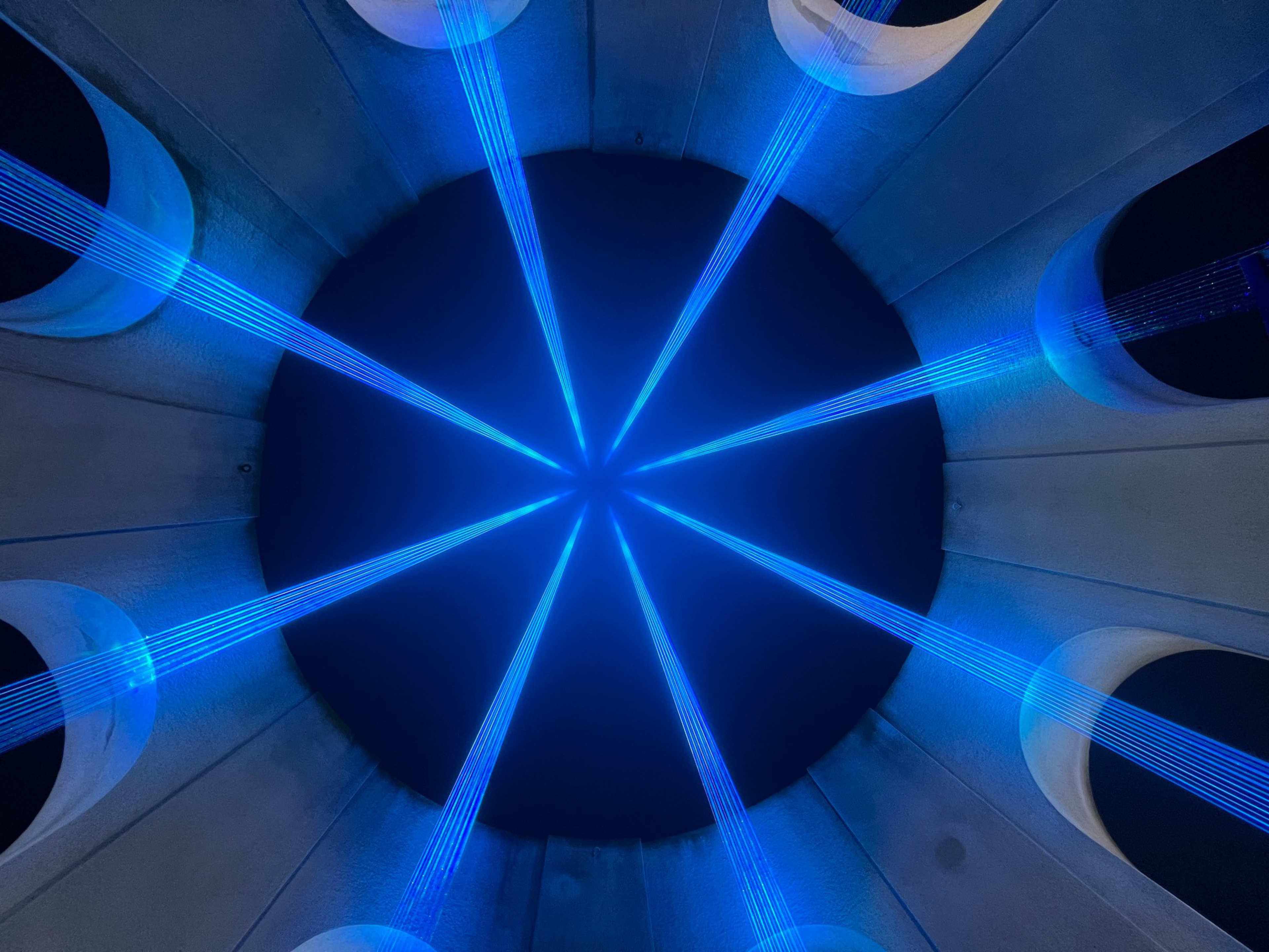 Blue laser beams radiate from the center of a dark circular space, framed by tall concrete pillars with circular cutouts.