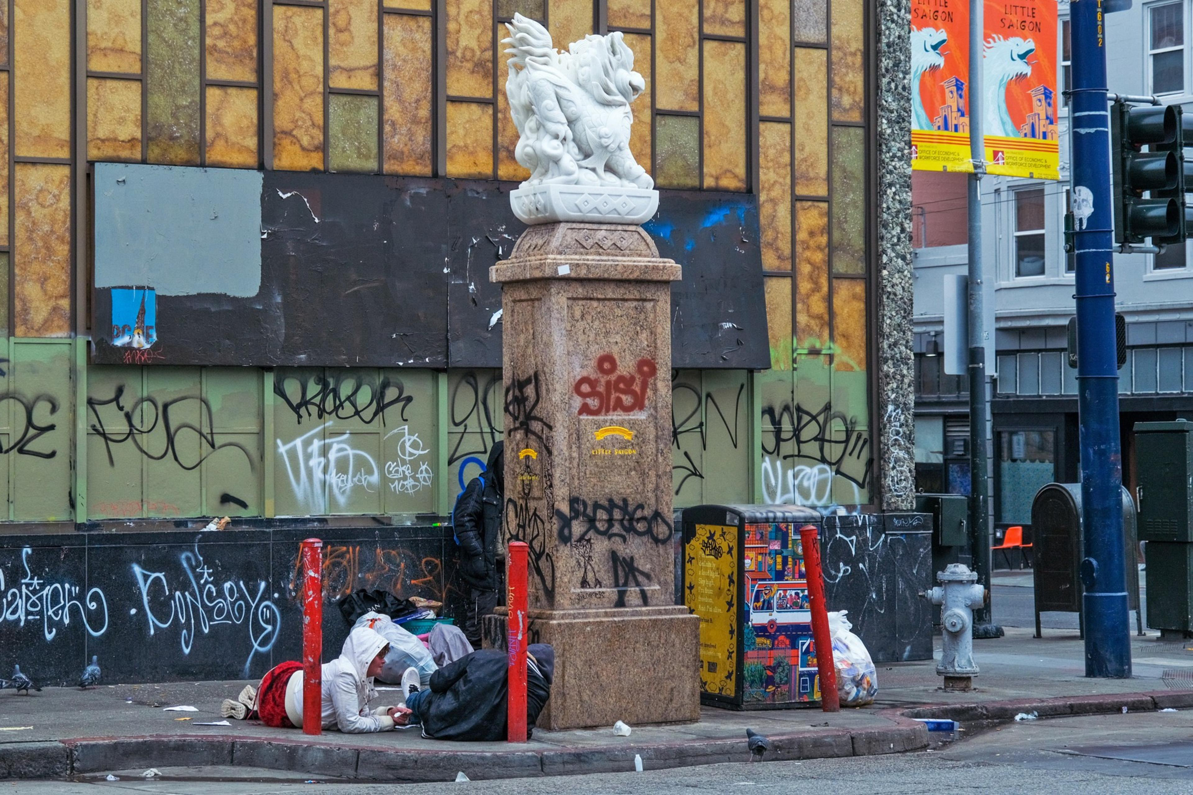 A graffiti-laden urban corner with a person resting by a statue base, surrounded by trash and discarded items.