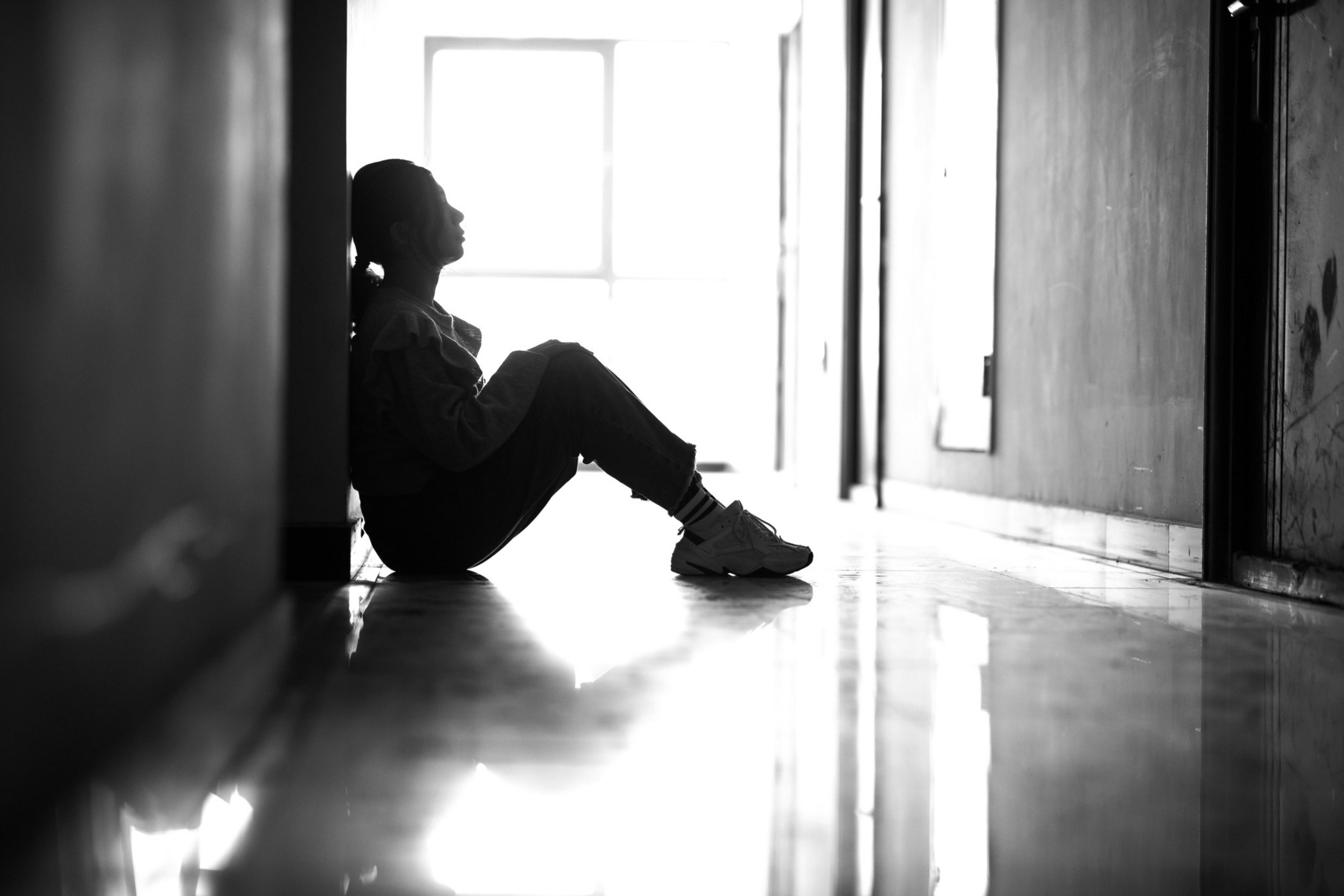 A person sits on the floor in a dimly lit hallway, leaning against a wall. They are in a reflective pose, with light shining through a window ahead.