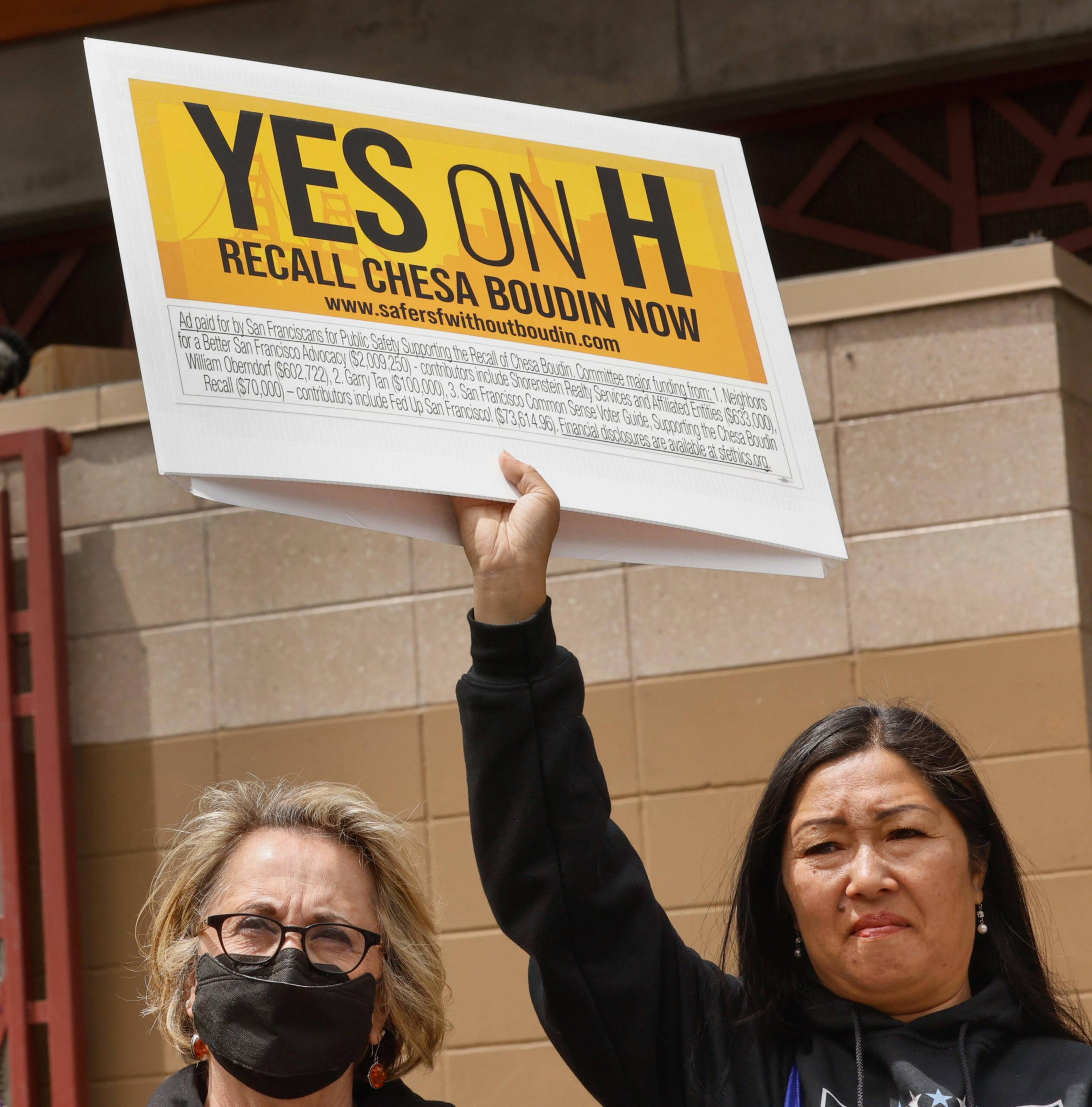 A woman holds a sign saying "YES ON H RECALL CHESA BOUDIN NOW" while standing next to another woman wearing glasses and a black face mask.