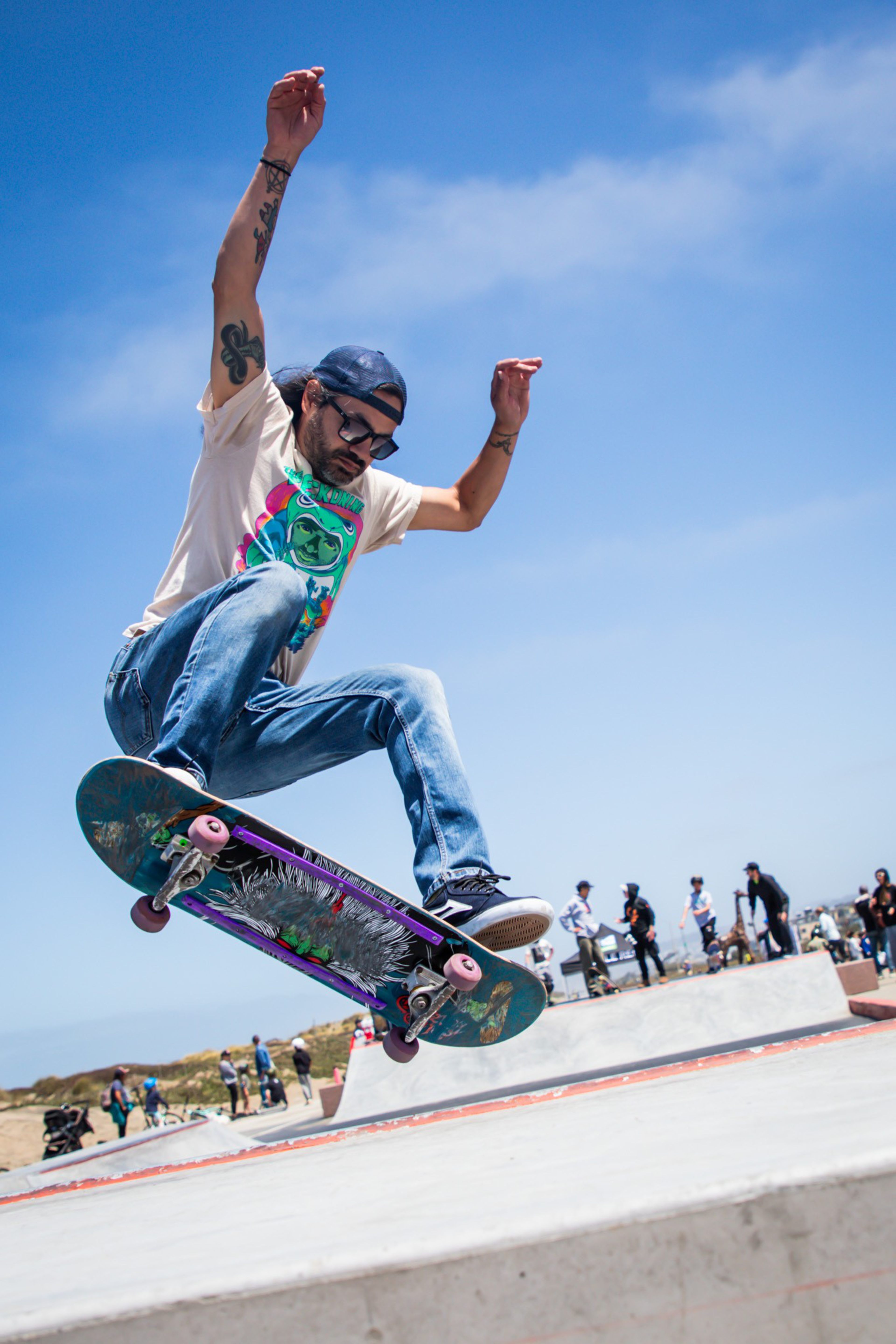 A skateboarder performs a trick mid-air, wearing a white t-shirt with a colorful design, jeans, and a cap. The background shows people at a skate park under a blue sky.