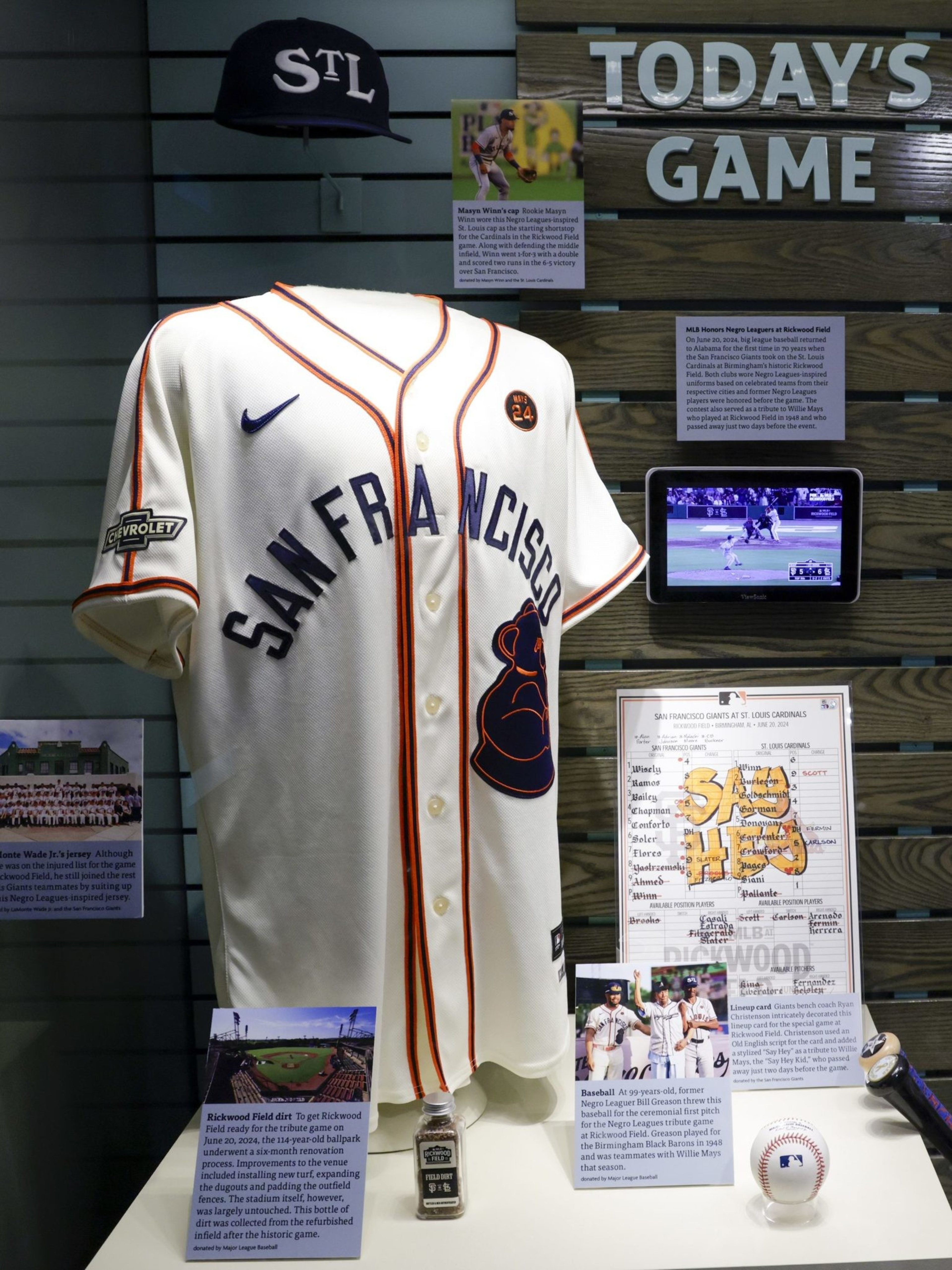 The image shows a San Francisco Giants jersey on display, surrounded by baseball memorabilia, including a cap, a lineup card, photos, and descriptive text panels.