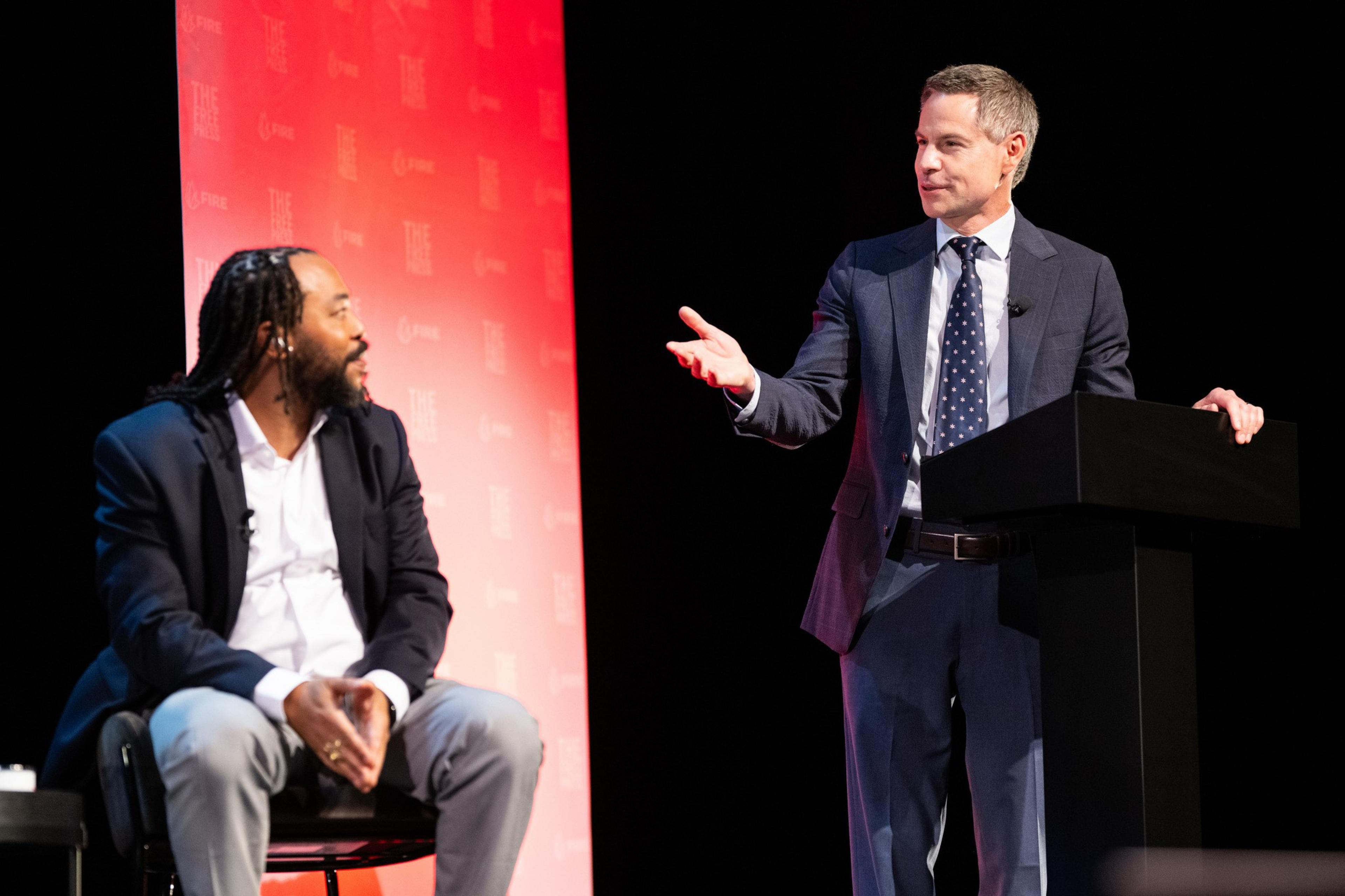A man is seated on the left, looking at another man in a suit standing at a podium. The background is red with subtle text patterns.