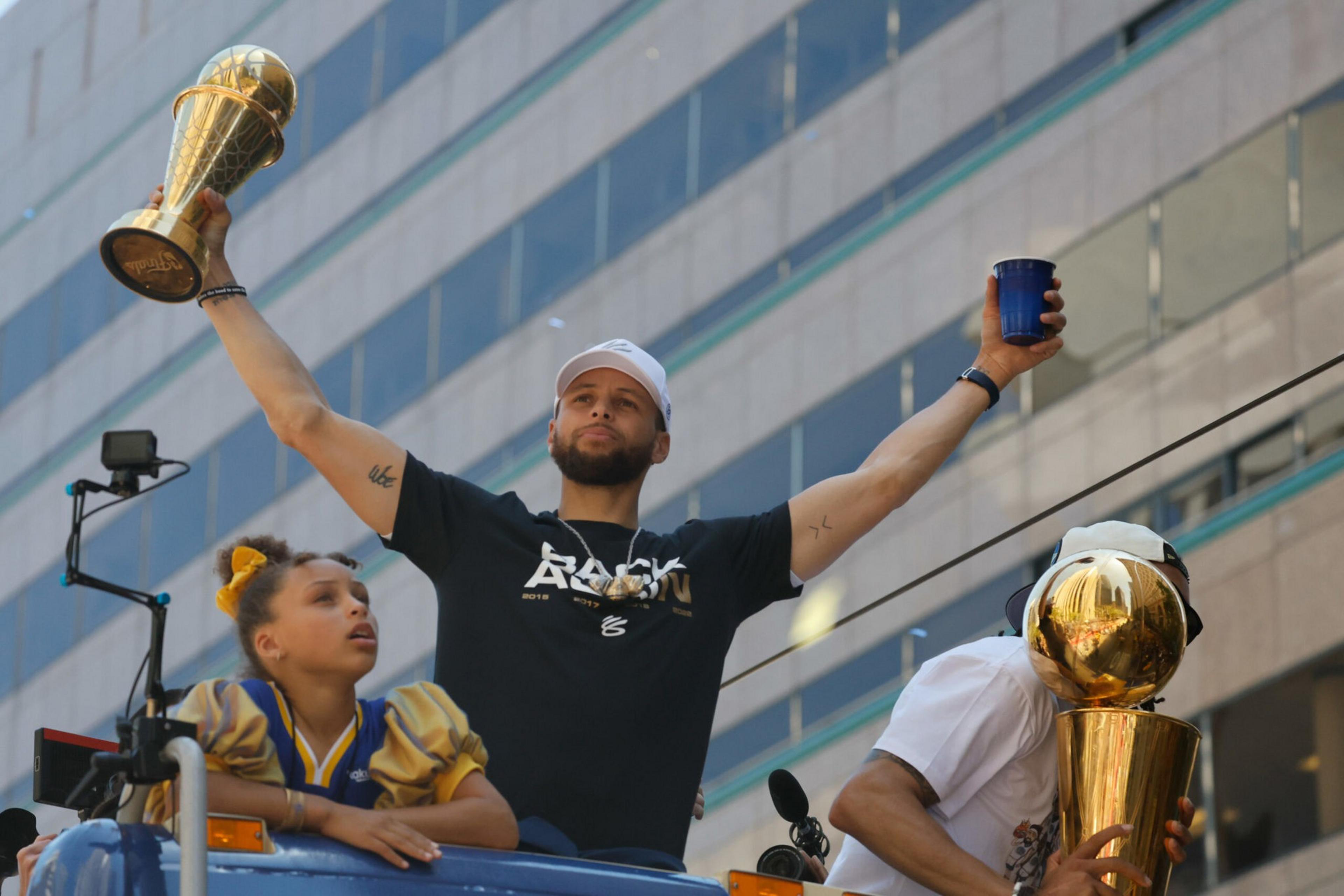 A man in a black shirt holds a trophy high with one hand and a cup in the other, while a girl in a sports jersey looks up beside him.