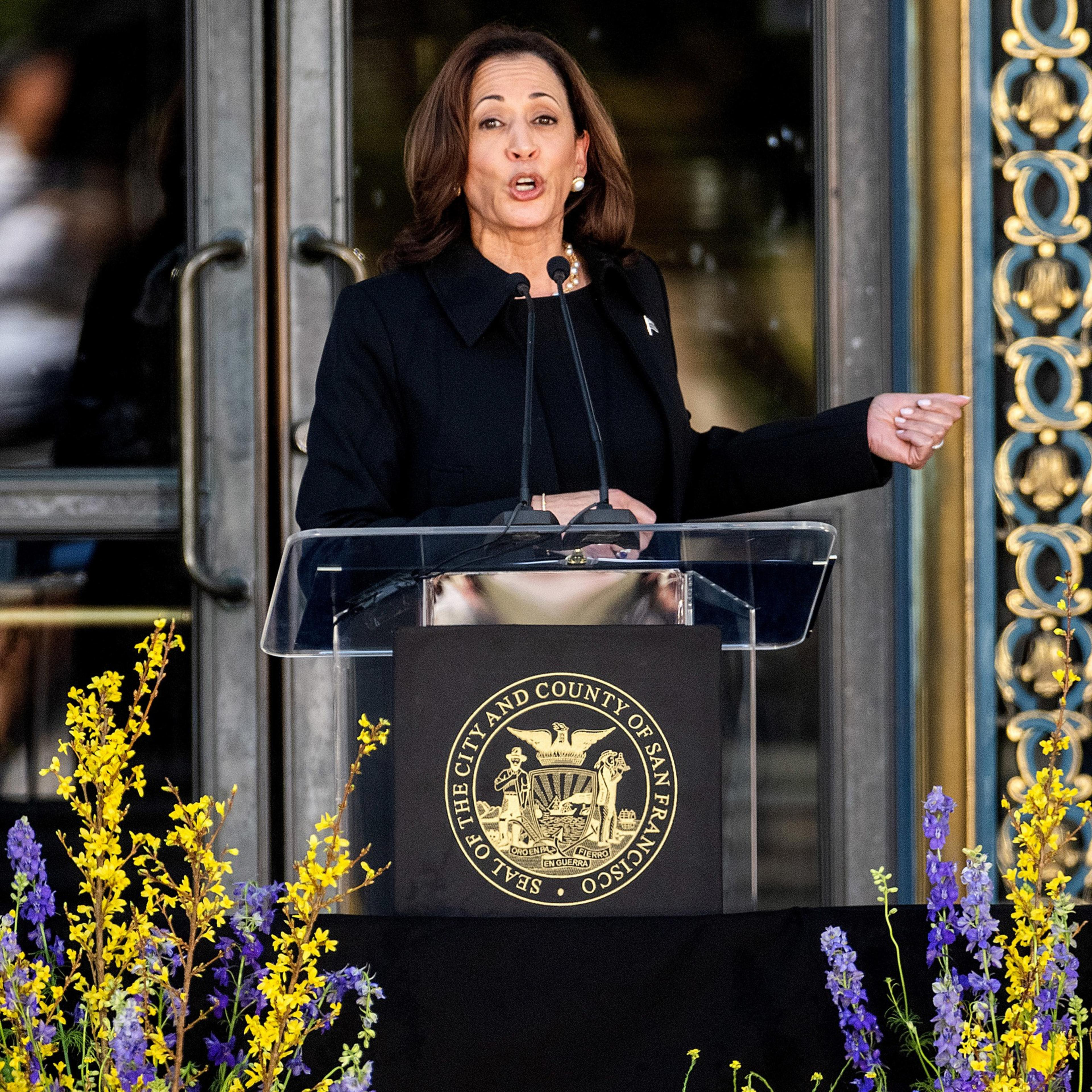 A woman in a black outfit speaks passionately at a podium adorned with the Seal of San Francisco, with colorful flowers in the foreground and ornate doors behind her.