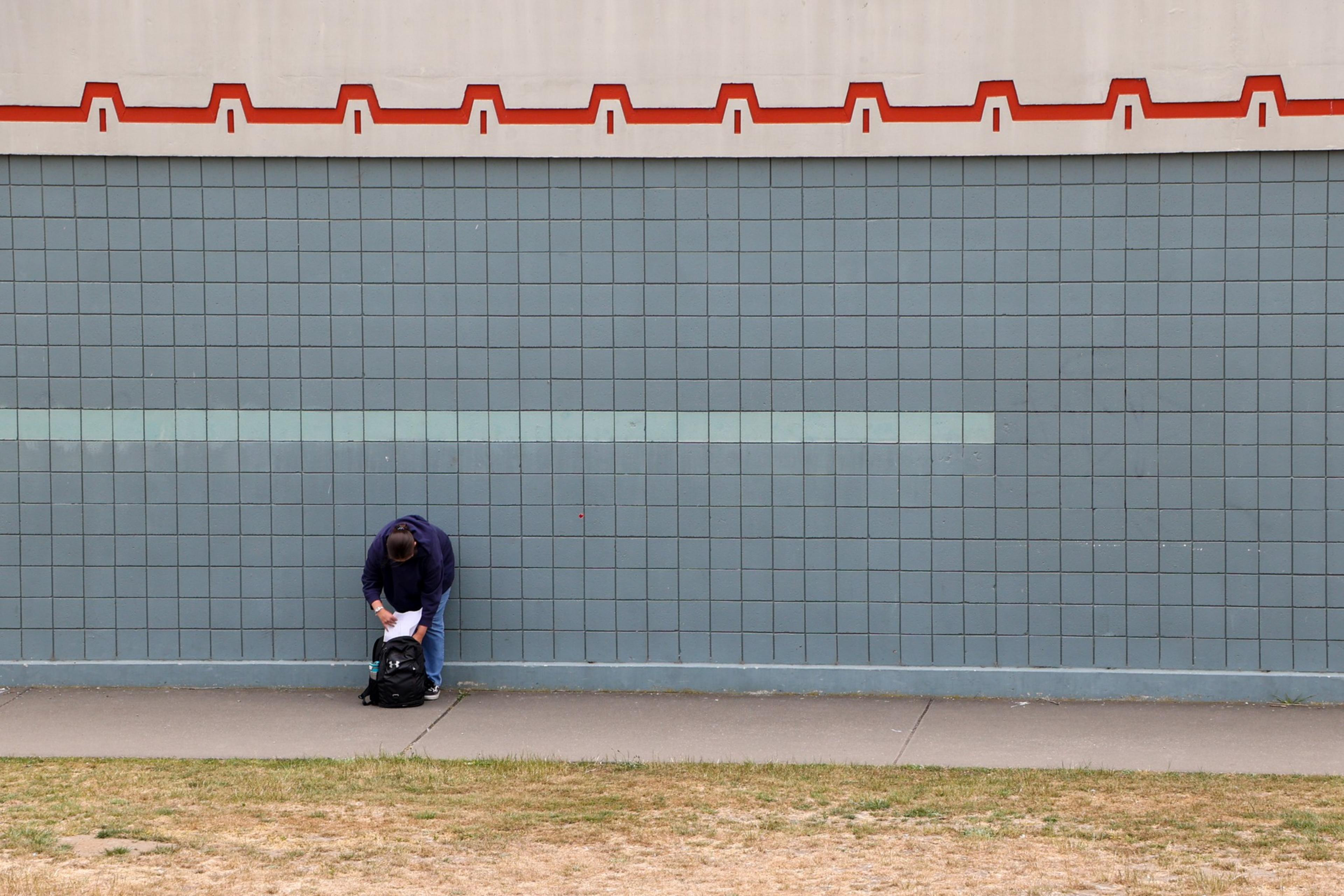 A person in a dark hoodie and jeans stands on a sidewalk, leaning over a black backpack, in front of a large blue-tiled wall with a beige section above.