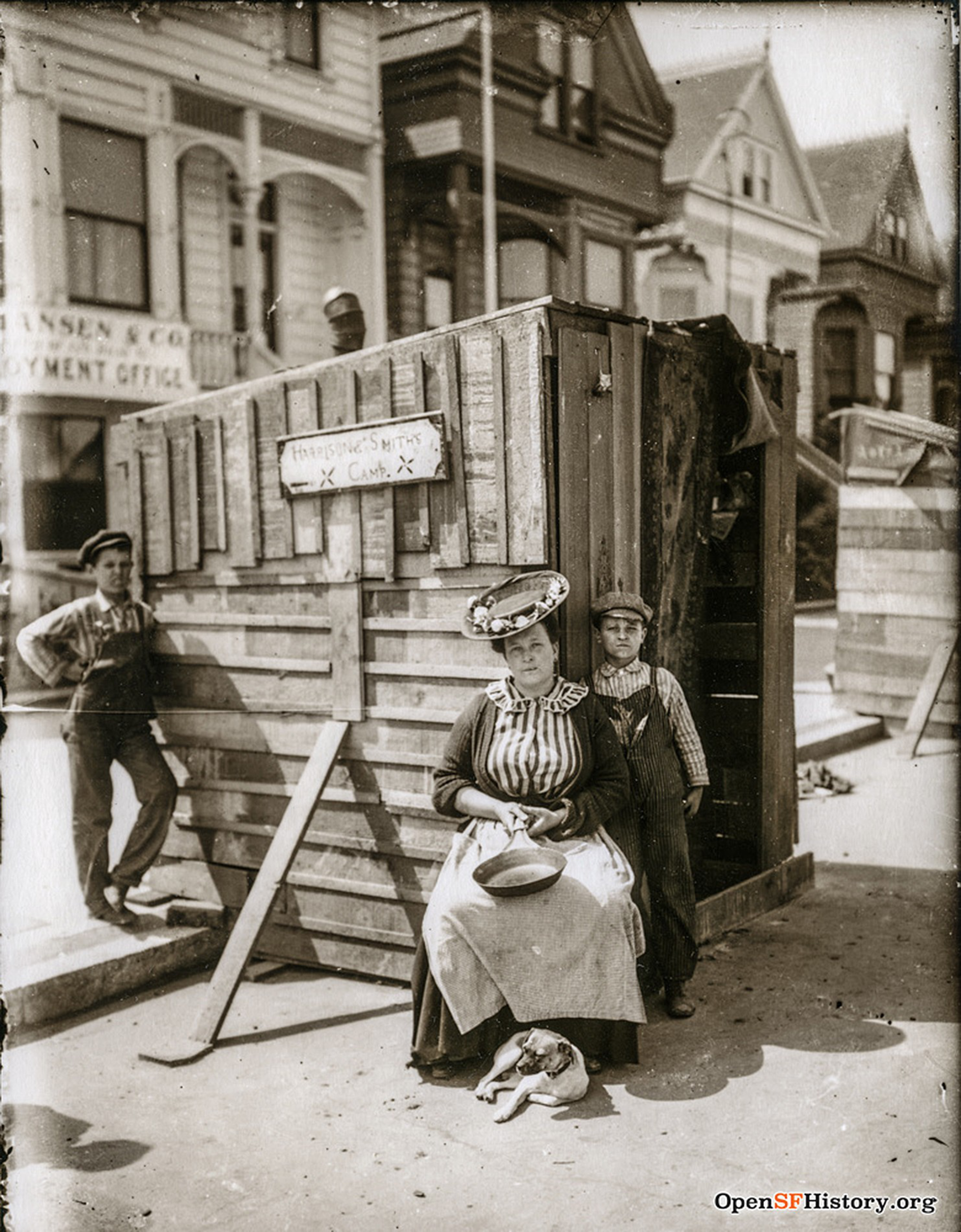 A woman in a striped dress and apron sits by a wooden structure labeled "Christian's Smith Camp," with two boys standing nearby and a small dog on the ground.