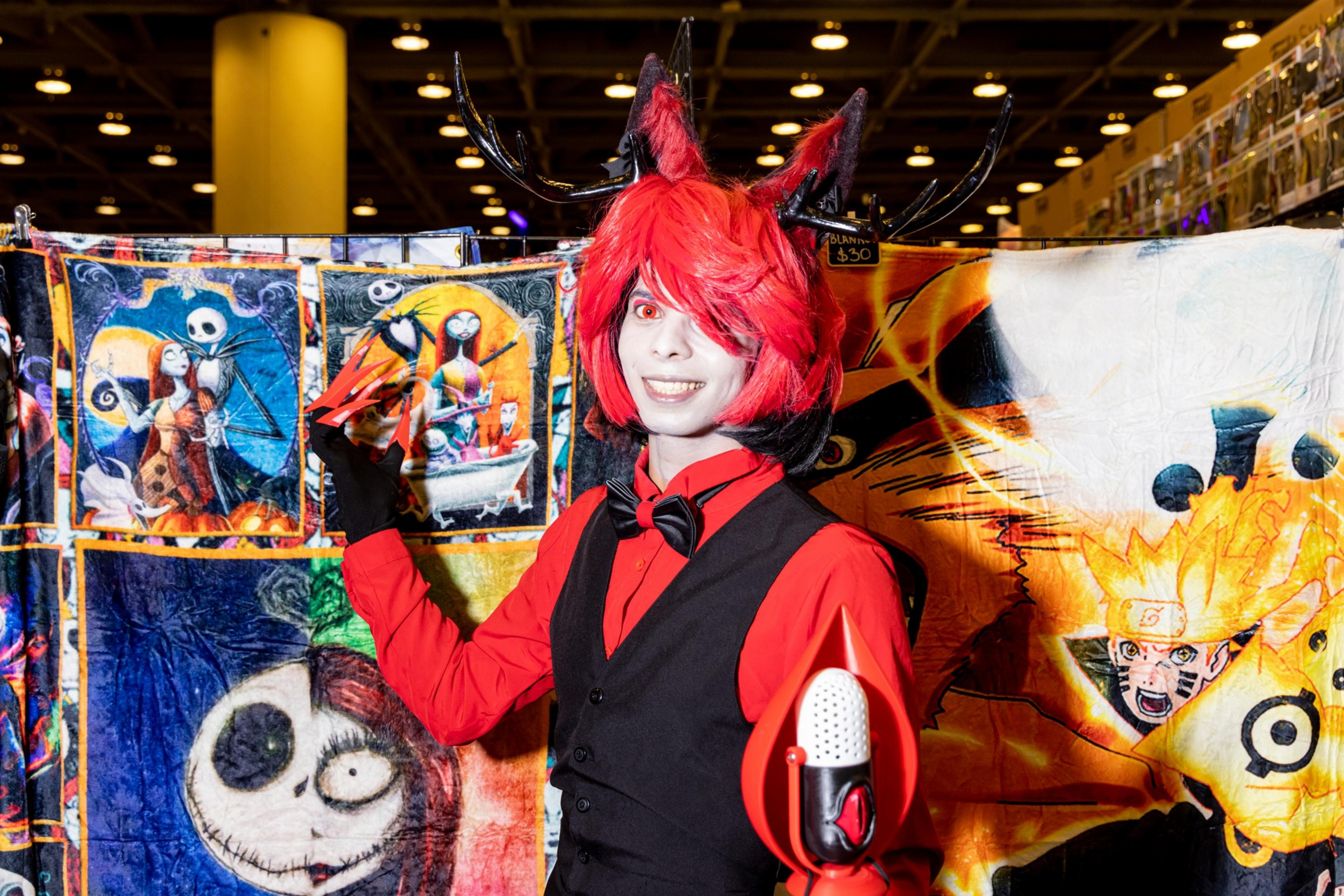 A person in a red and black costume with red hair and pointed ears stands in front of colorful, character-themed blankets, holding up a claw-like prop.