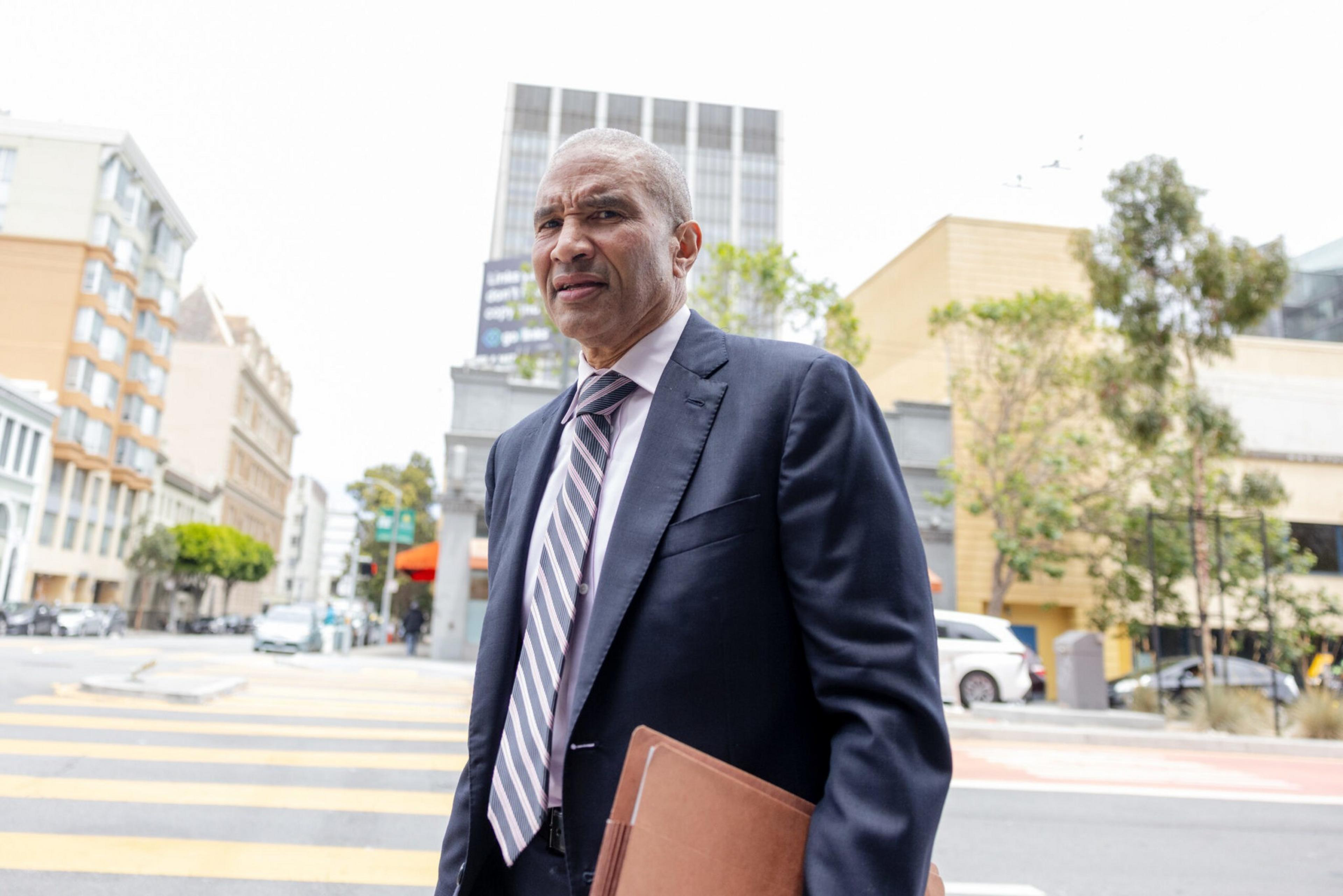 A man in a suit with a briefcase walks on a city street, with buildings and pedestrian crossing behind him.