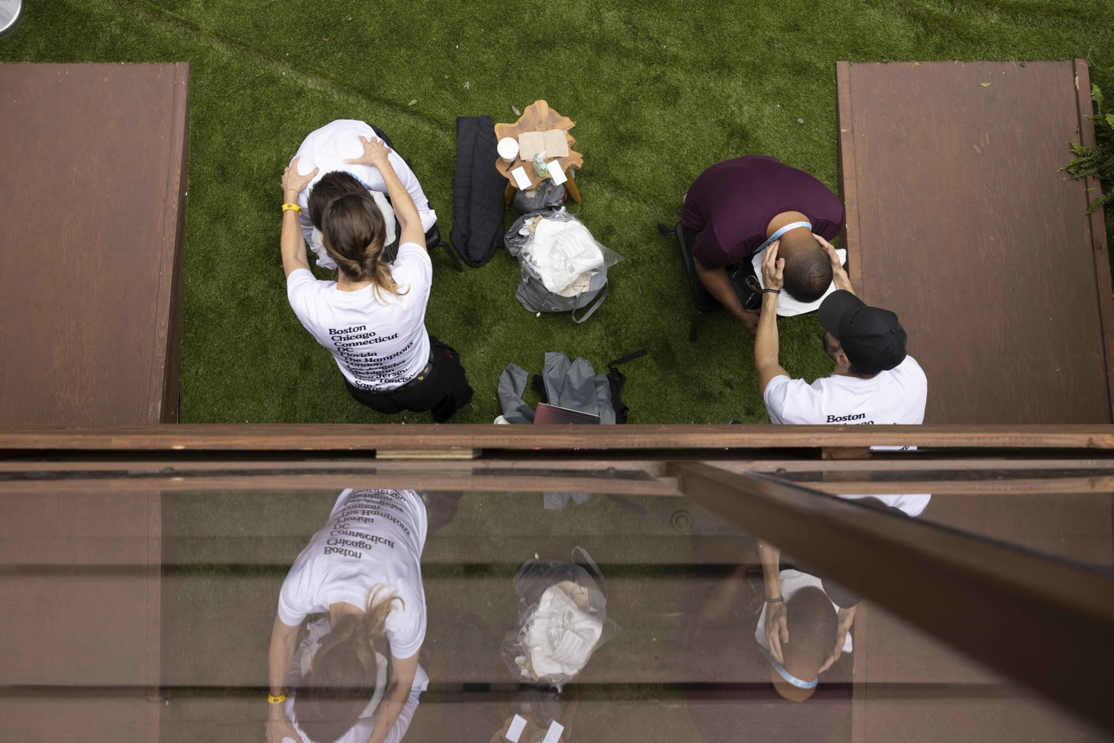 Two people are receiving massages on a grassy area, with two others standing behind them. A small wooden table with various items is placed between them.