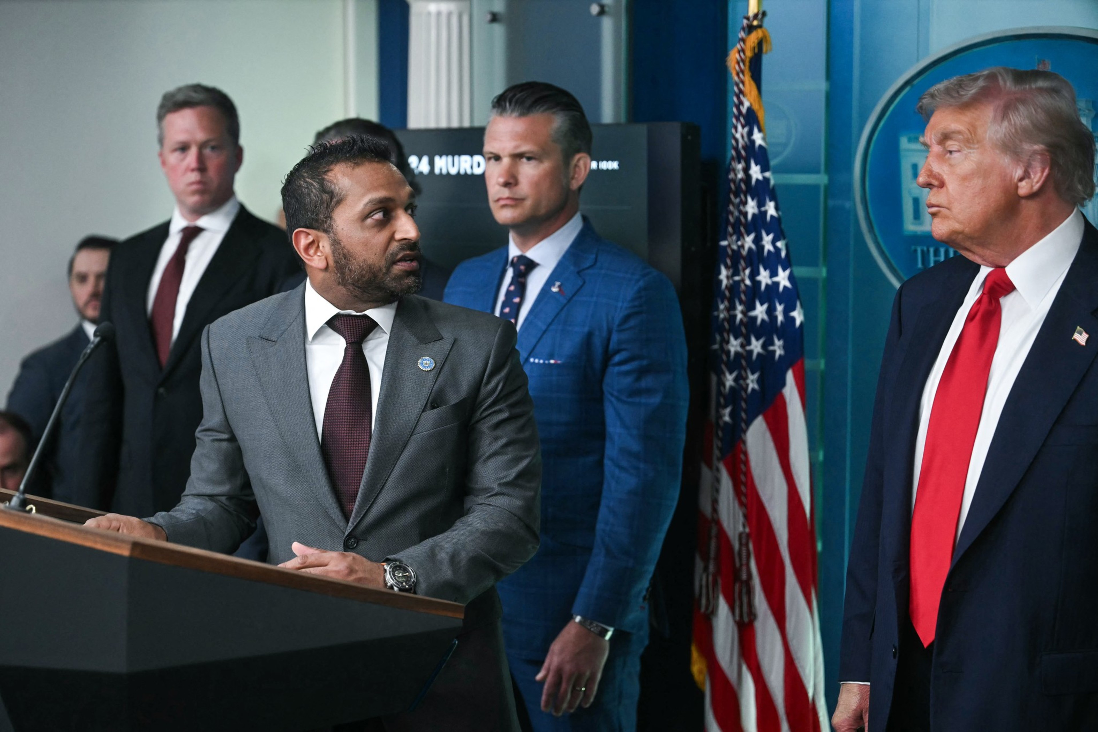 A man in a gray suit speaks at a podium while three men in suits, including one with an American flag pin, listen attentively nearby.