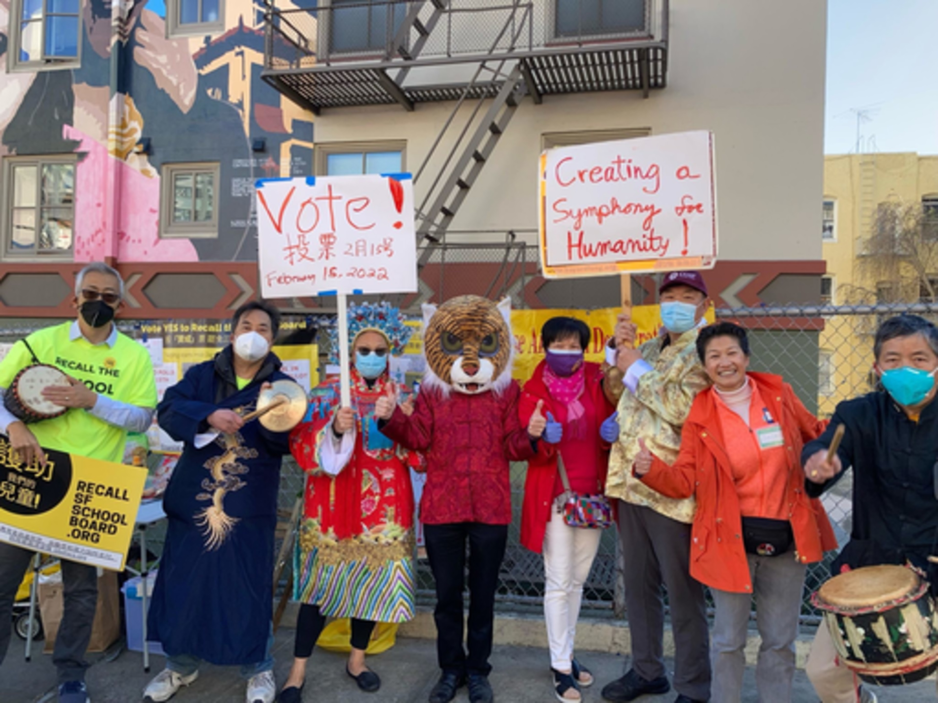 A group of people, some in traditional attire and wearing masks, hold signs urging to vote on February 15, 2022, and promote humanity, with one wearing a tiger mask.