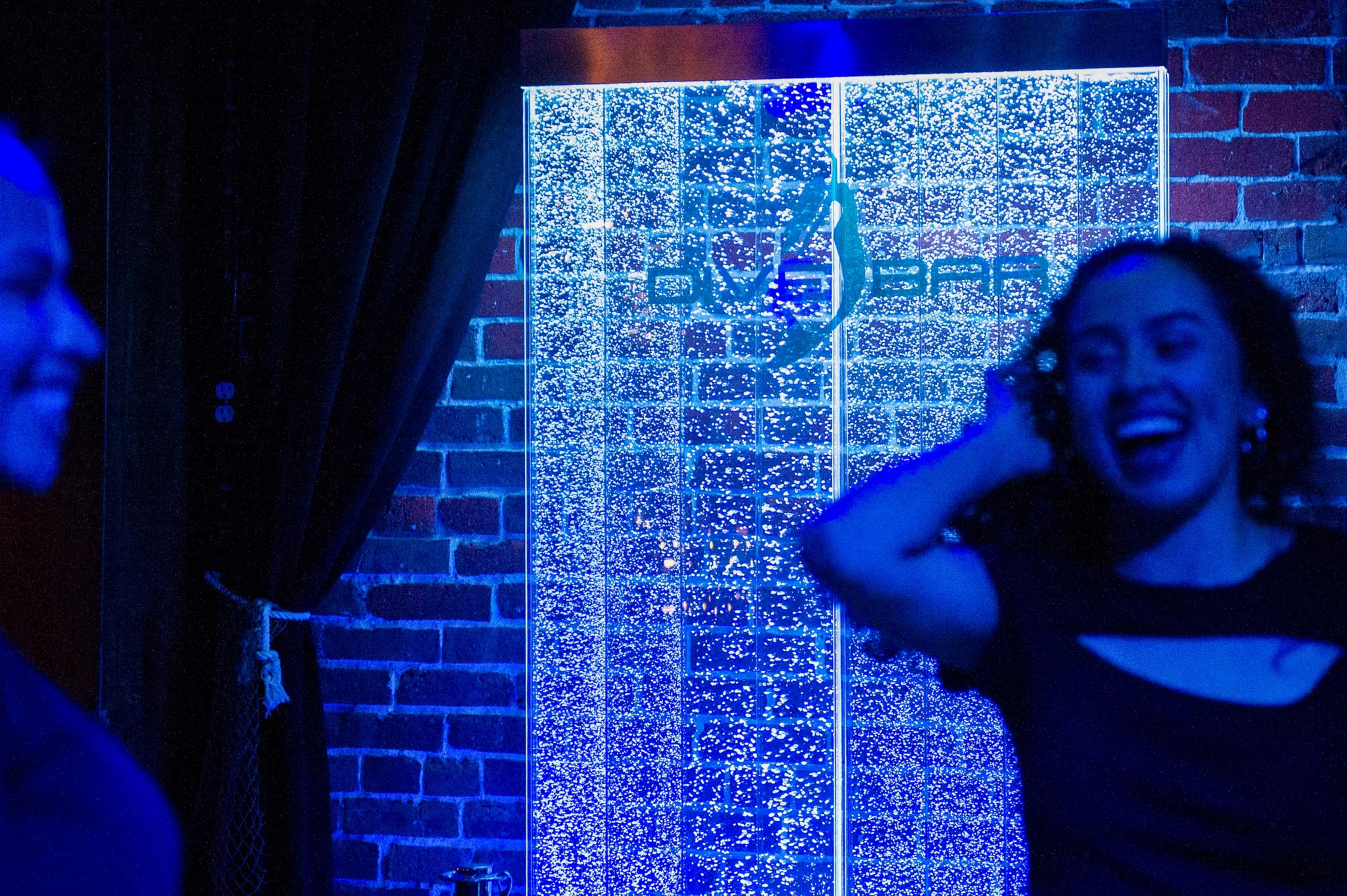 Two women smile and talk by a wall with a glowing blue vertical panel filled with sparkling bubbles and a faint pole silhouette.