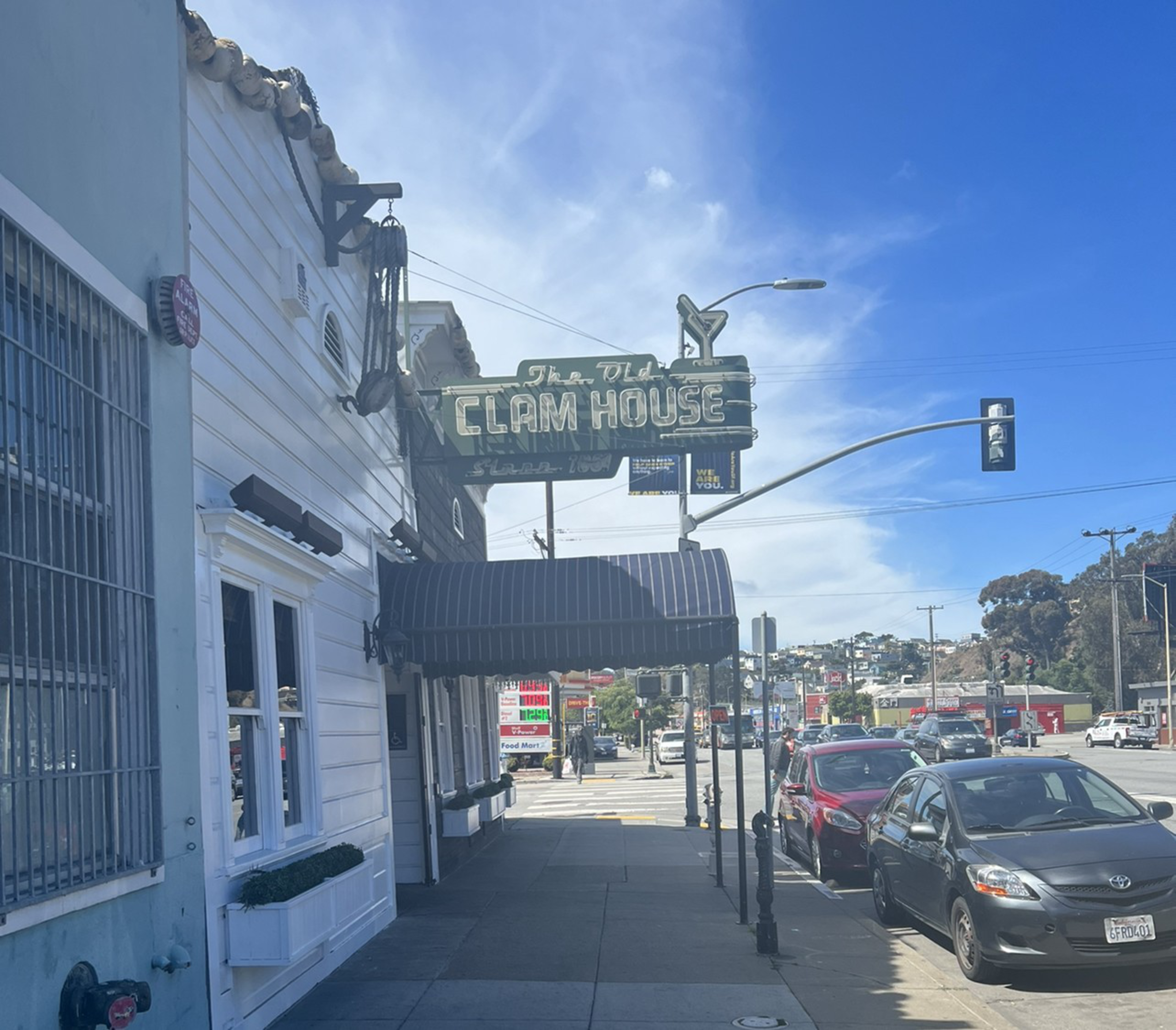 A street view of The Old Clam House restaurant with a green vintage sign, striped awning, parked cars, and a clear blue sky.