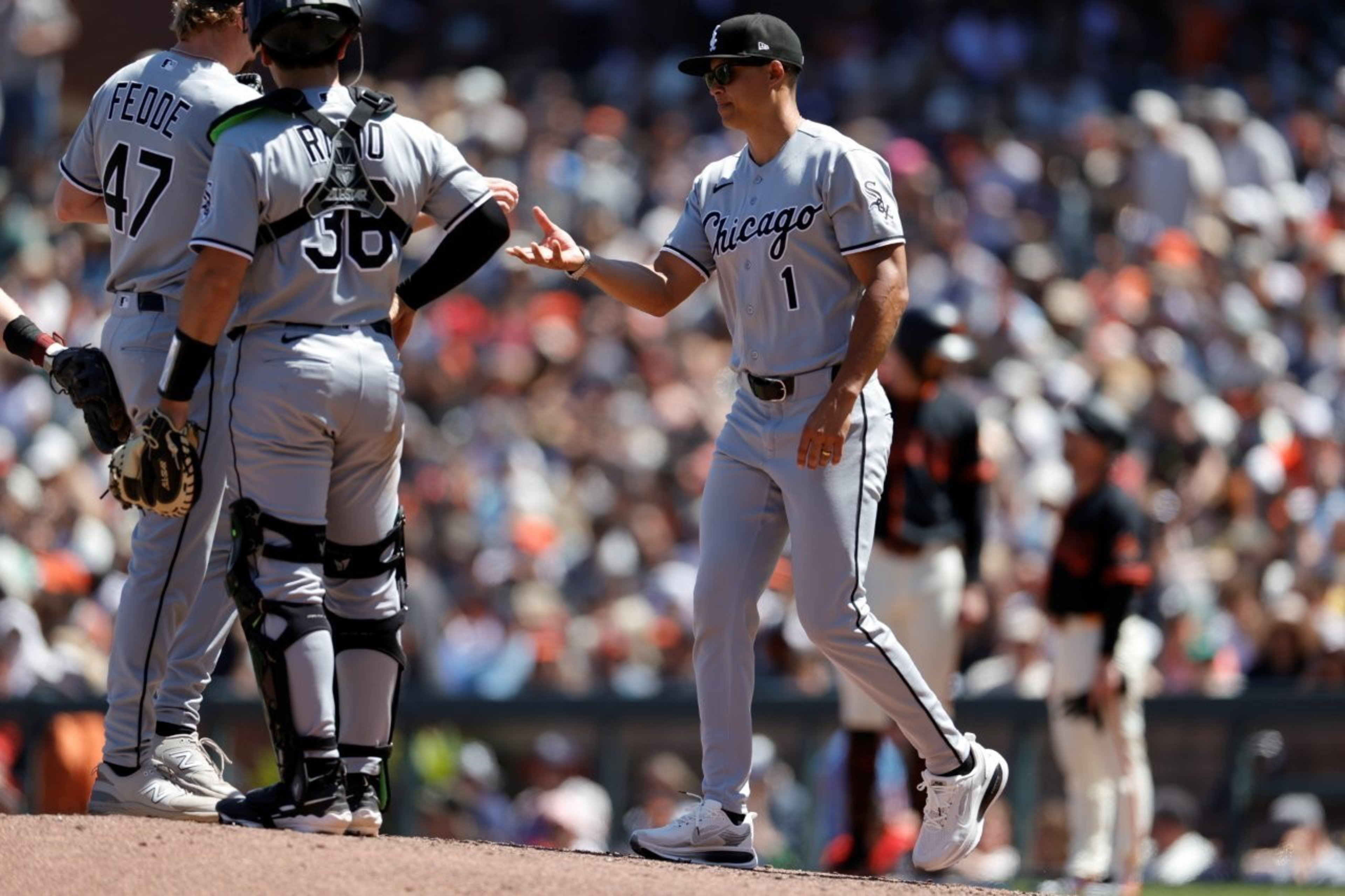Three Chicago White Sox players, including a pitcher and catcher, meet on the mound during a sunny baseball game against a blurred opposing team.
