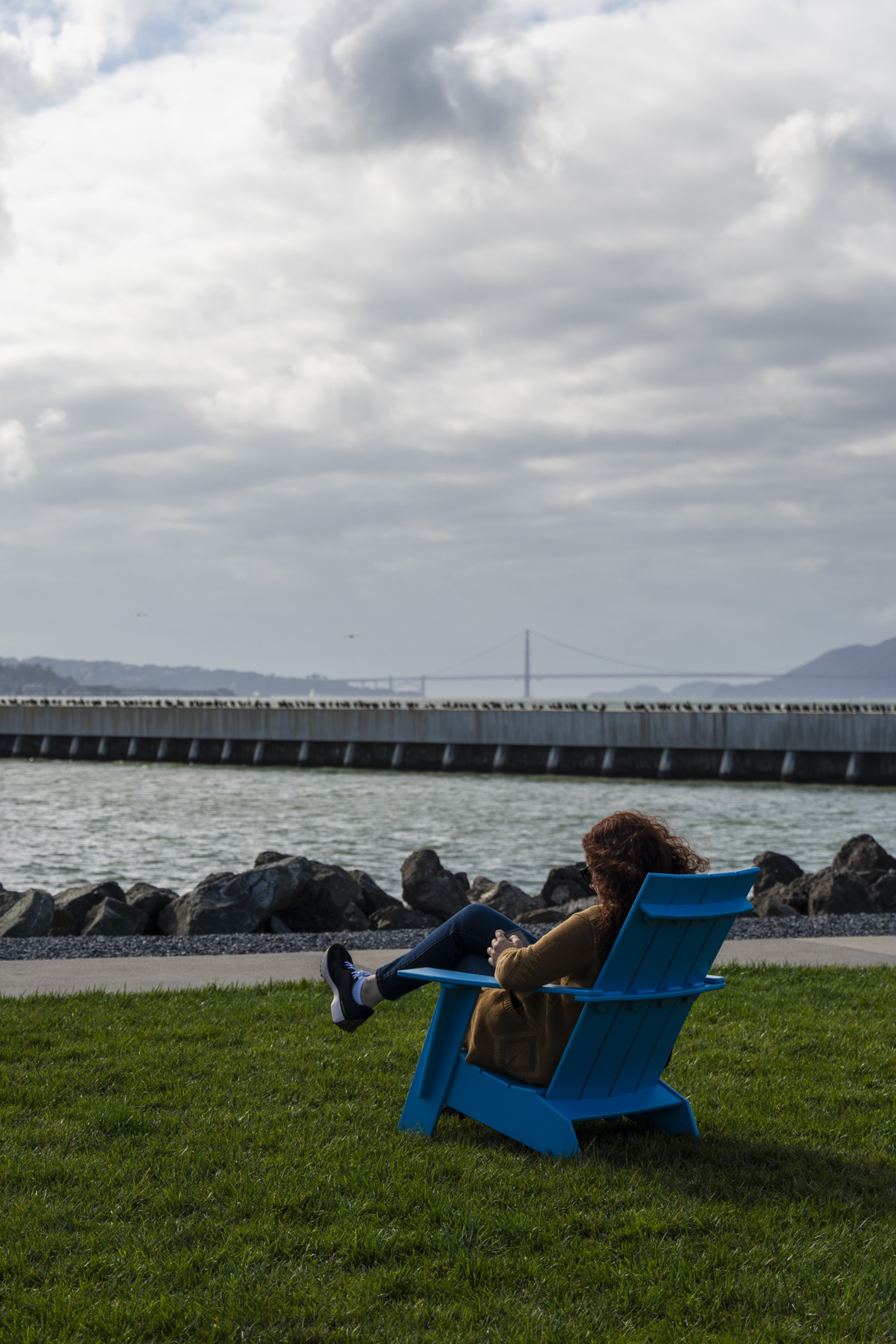 A person with curly hair sits in a blue Adirondack chair on green grass, facing water with rocks and a distant bridge under a cloudy sky.