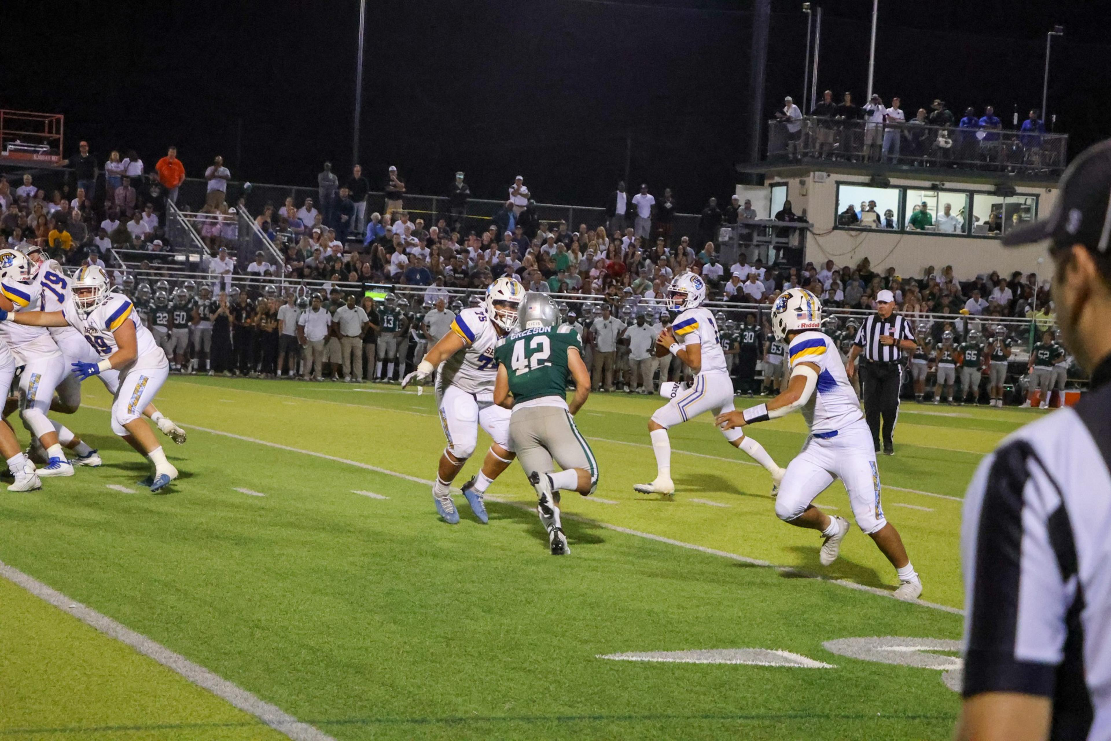 A football player in a green jersey runs toward the quarterback, who is preparing to throw the ball as teammates in white jerseys block.