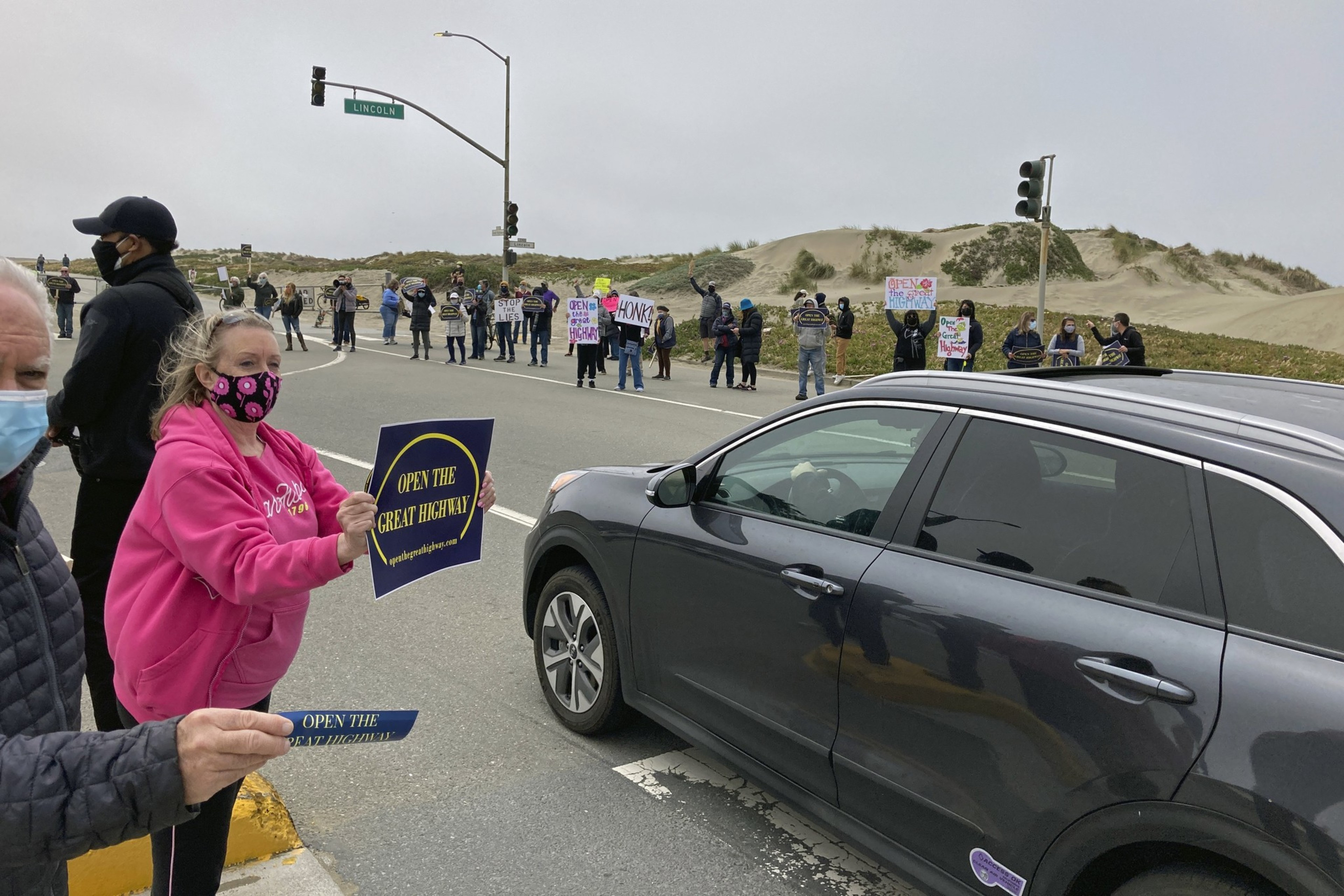 People are gathered at an intersection with signs protesting to &quot;Open the Great Highway,&quot; some standing on the roadside and others in a line across the street.