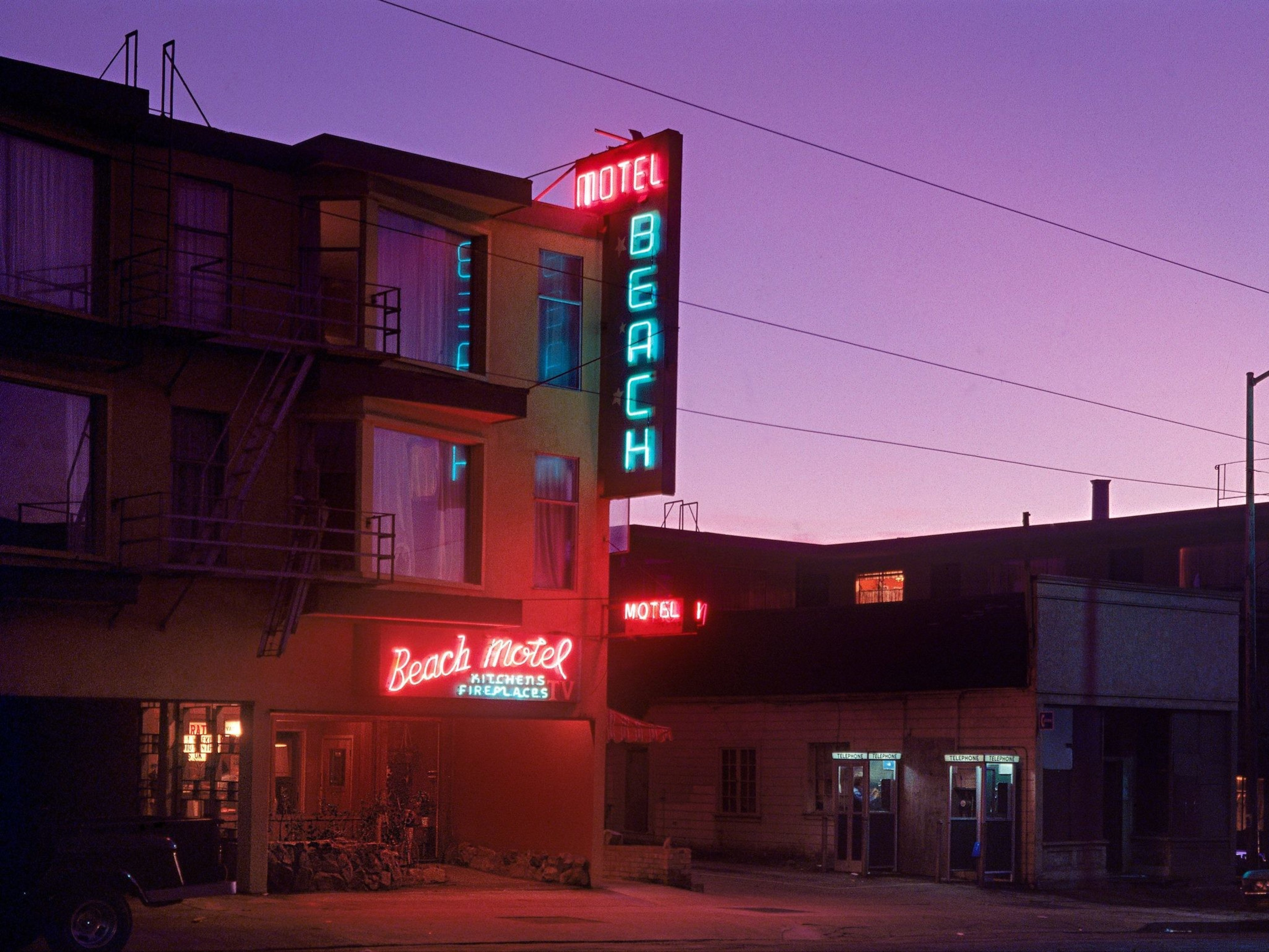 The image shows a vintage motel with neon signs reading &quot;Beach Motel,&quot; &quot;Kitchens,&quot; and &quot;Fireplaces.&quot; It's dusk, casting a purple hue over the scene.