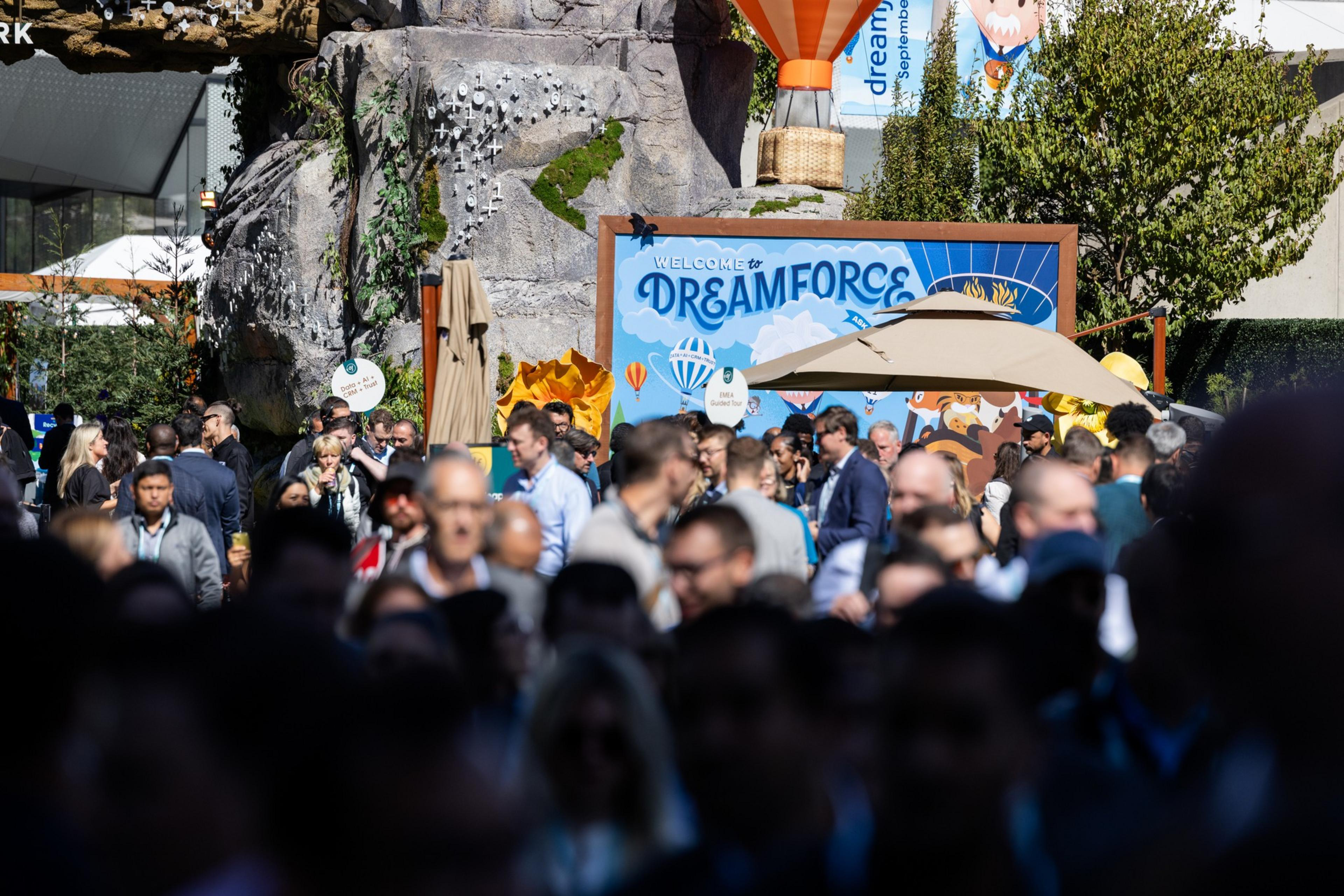 A large crowd of people gathers outdoors near a colorful "Welcome to Dreamforce" sign with a hot air balloon decoration above it.