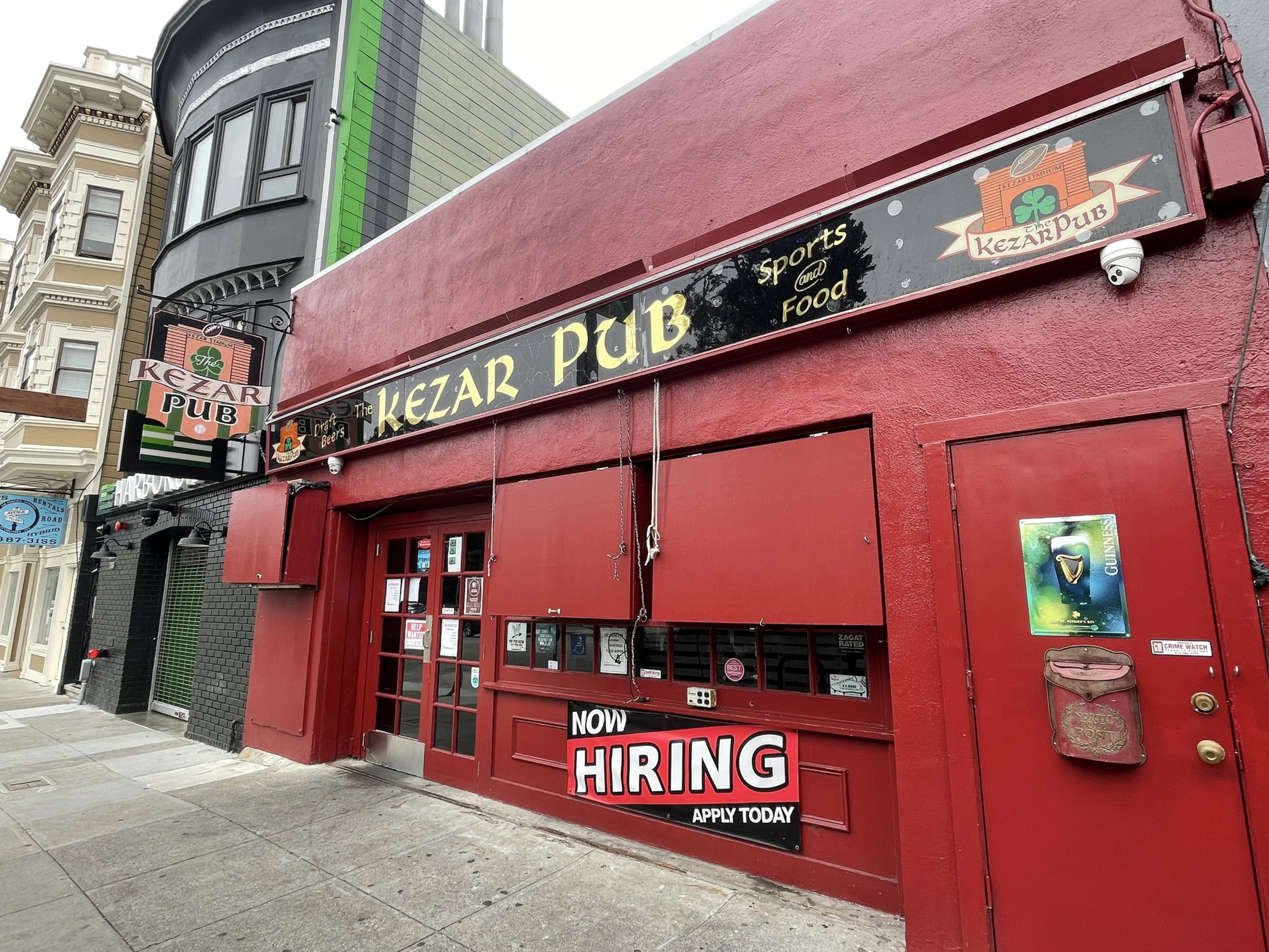 A red-painted pub named Kezar Pub with signs advertising sports, food, and a “Now Hiring” banner is located on a city sidewalk.