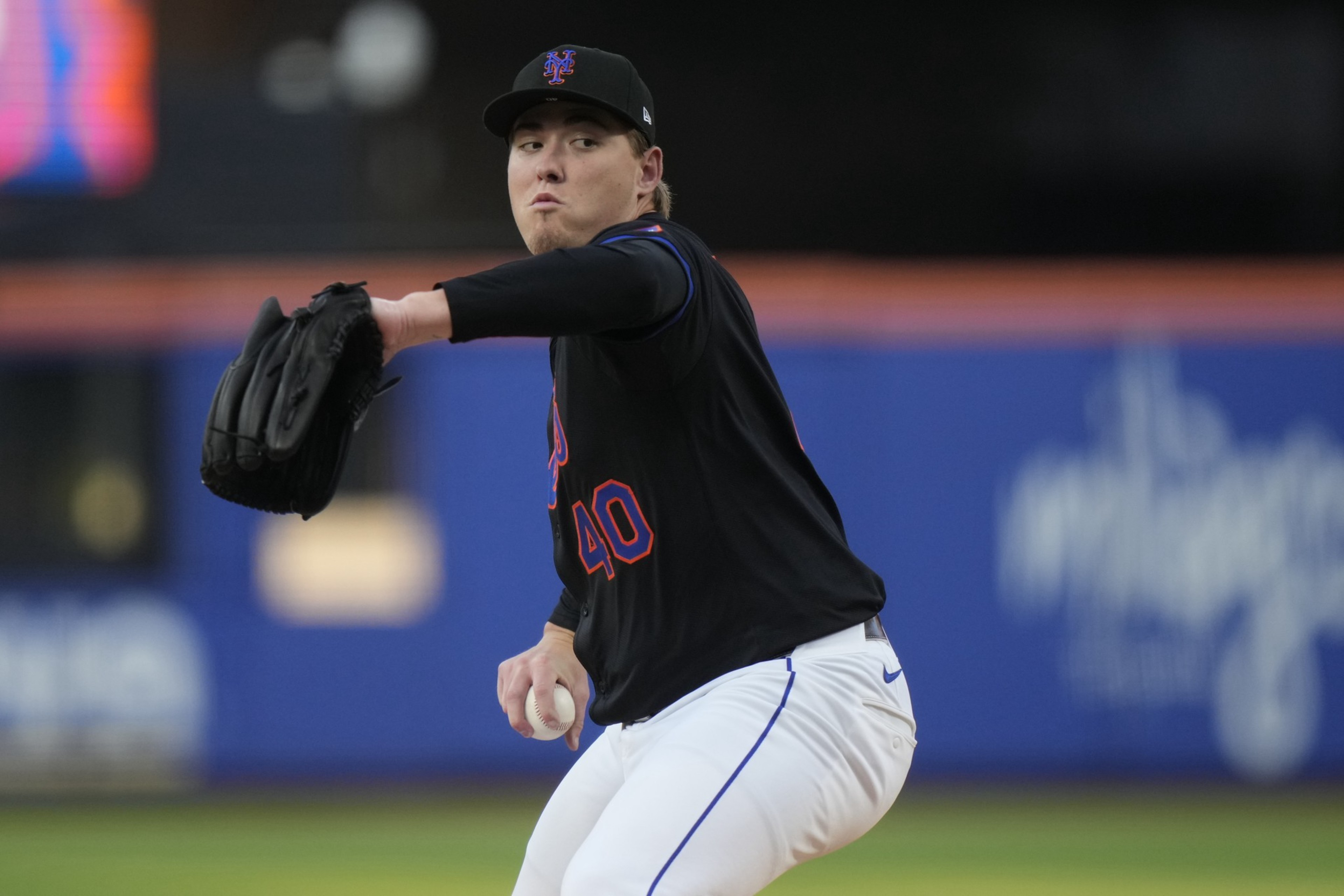 A baseball pitcher wearing a black jersey with number 40 is mid-throw, gripping a baseball in his right hand and wearing a black glove on his left.
