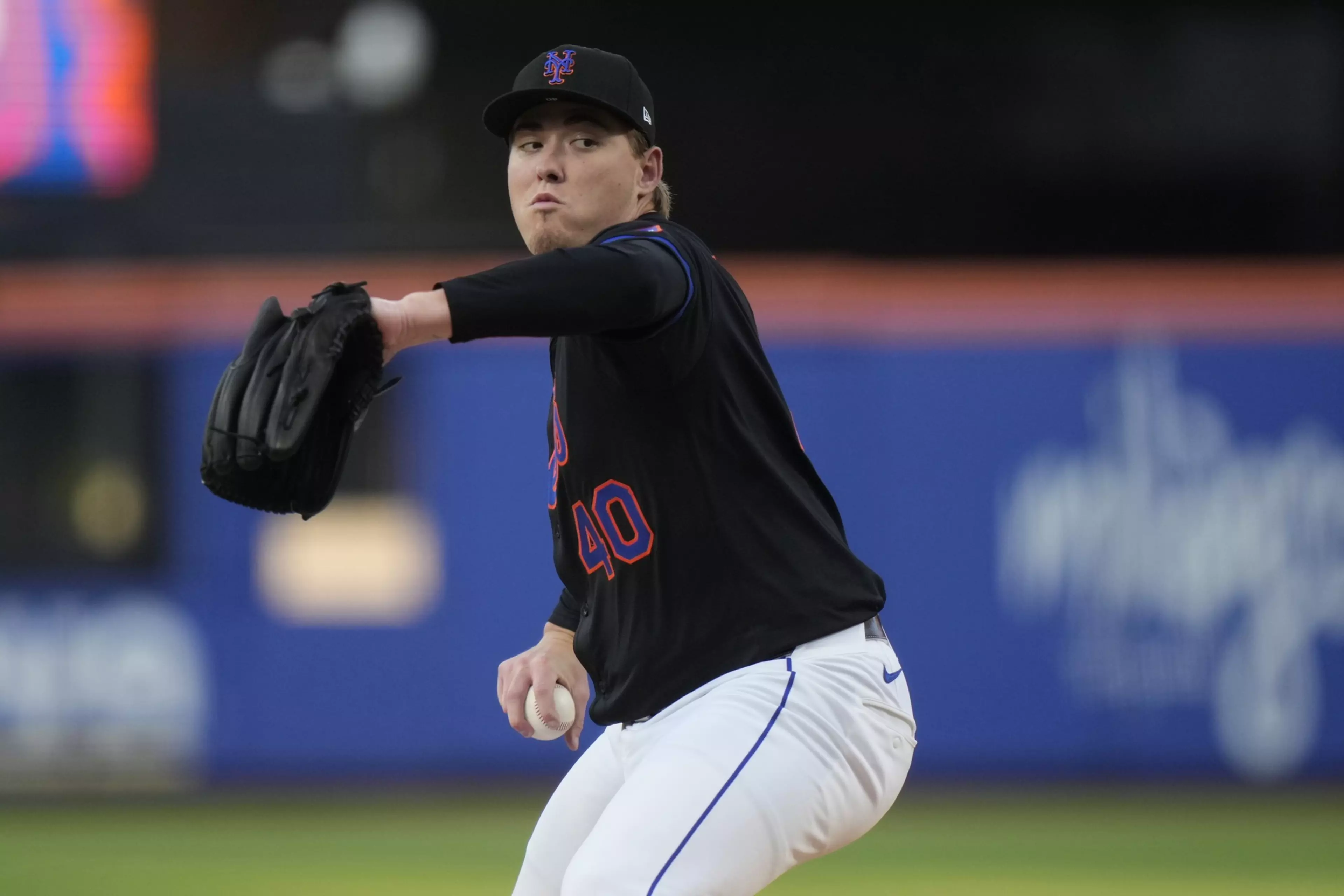 A baseball pitcher wearing a black jersey with number 40 is mid-throw, gripping a baseball in his right hand and wearing a black glove on his left.