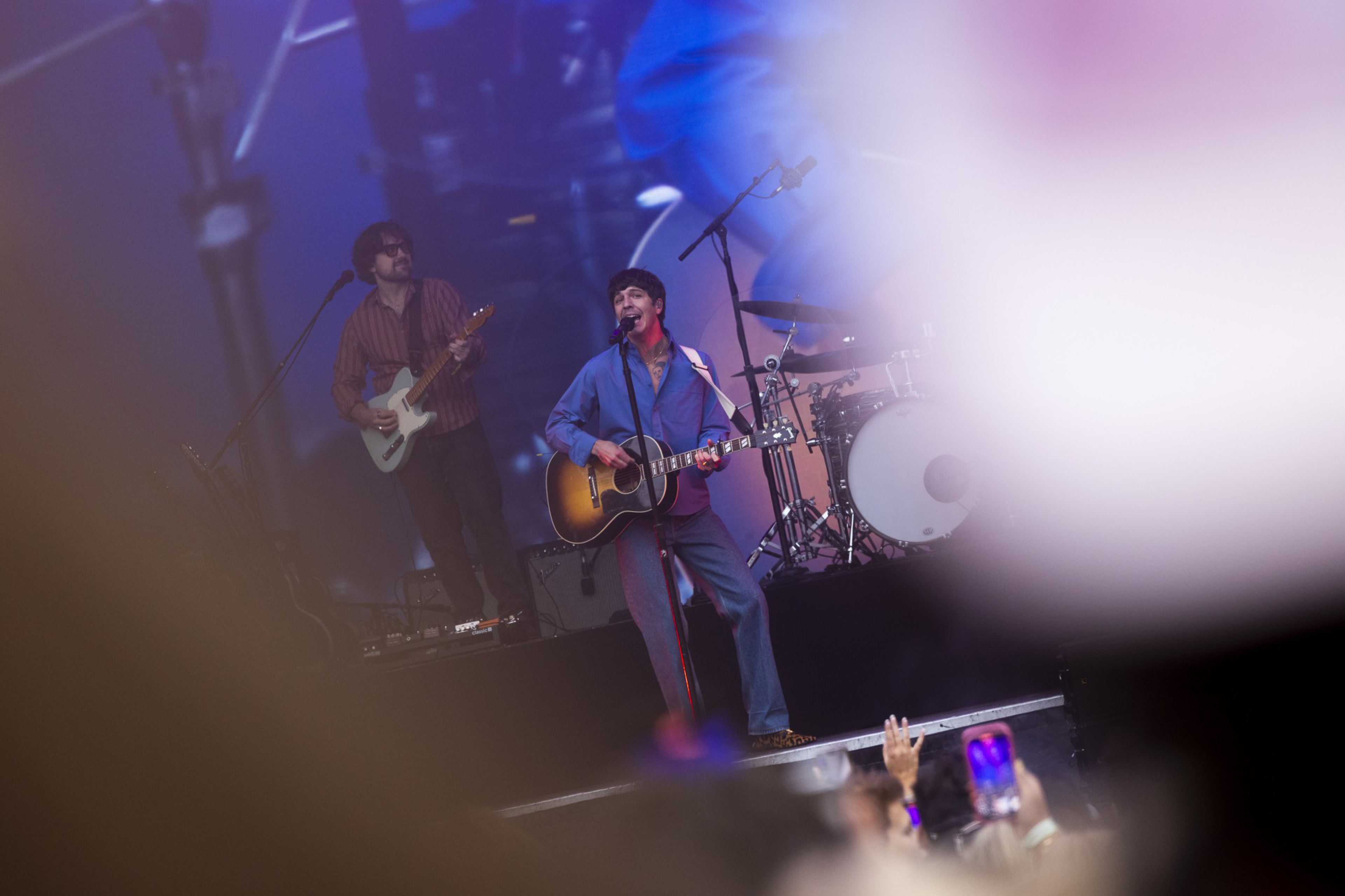 A musician in a blue outfit sings and plays an acoustic guitar on stage while another person plays electric guitar behind him.