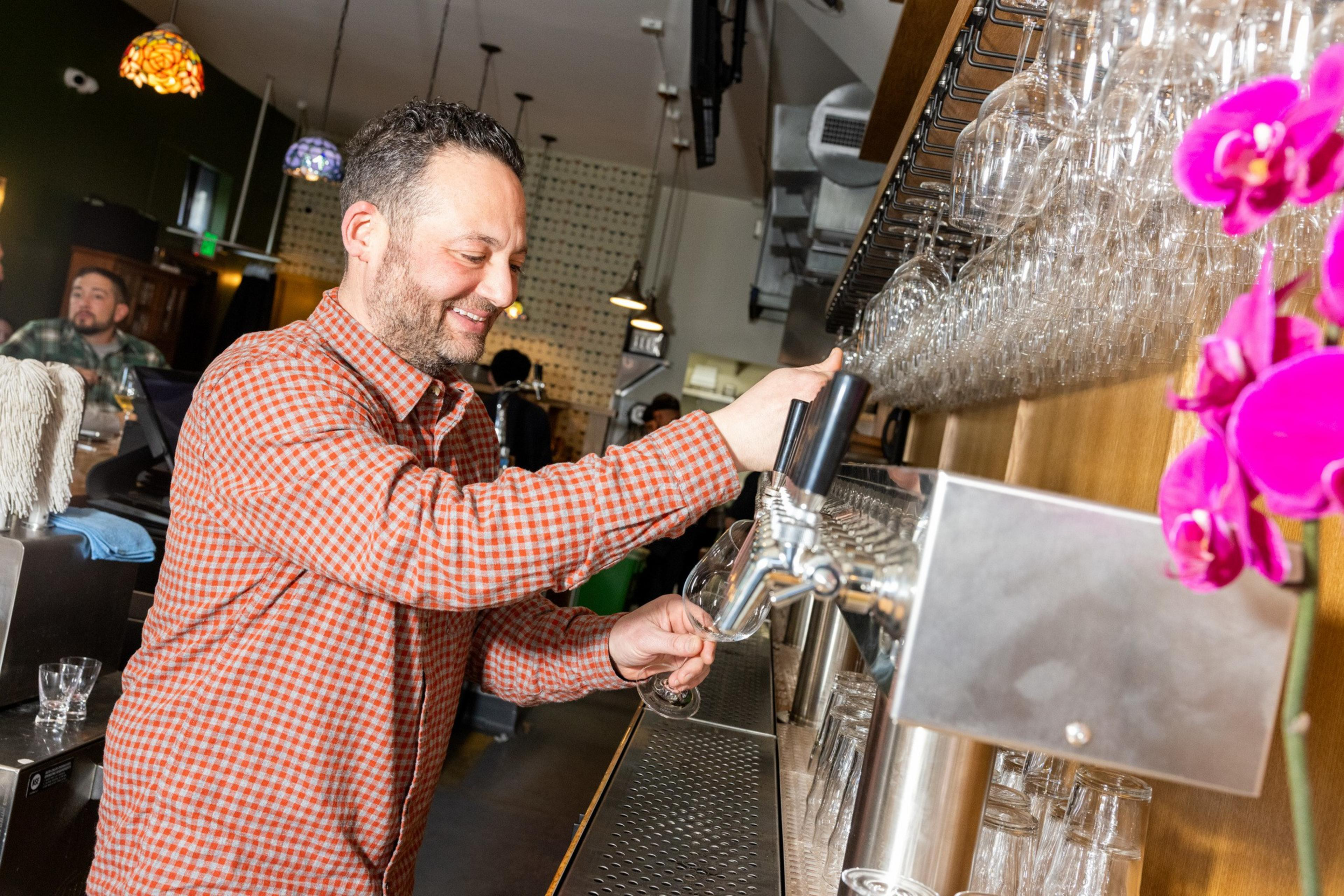 A man in a checkered shirt is smiling while pouring a drink from a tap behind a bar. Glassware is neatly stacked above, and pink flowers are nearby.