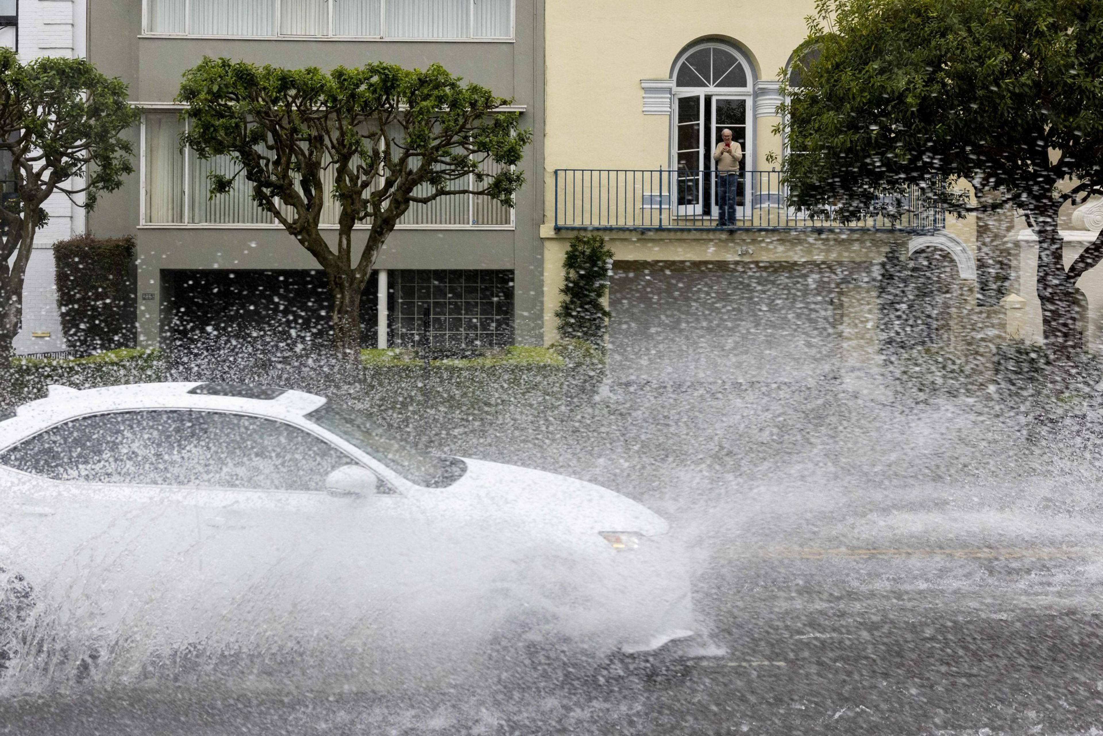 A white car speeds through a large puddle, creating a big splash of water. A person observes the scene from a balcony in front of a building with trees nearby.