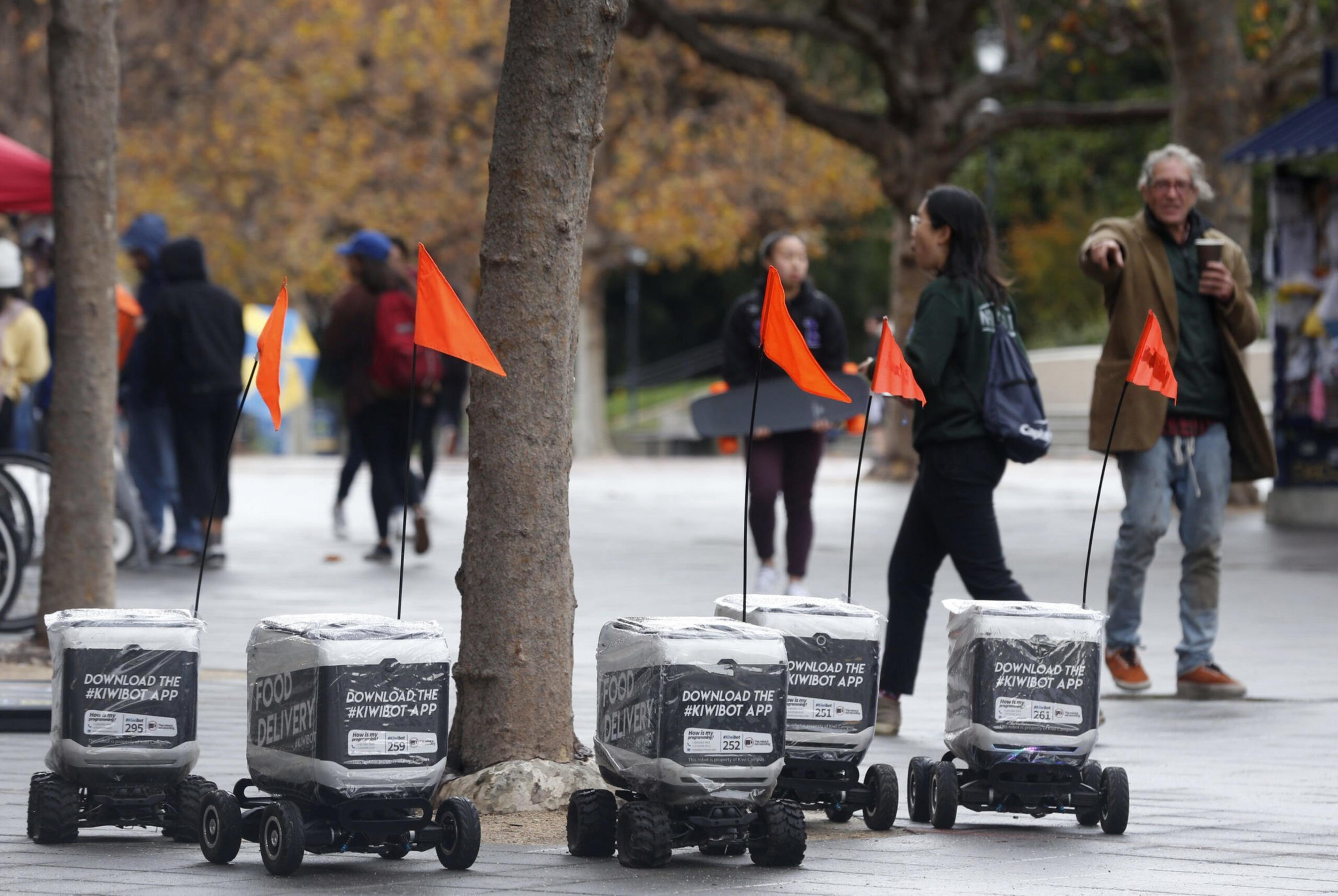 Five small autonomous food delivery robots with orange flags are lined up on a sidewalk, while people walk and stand in the background.