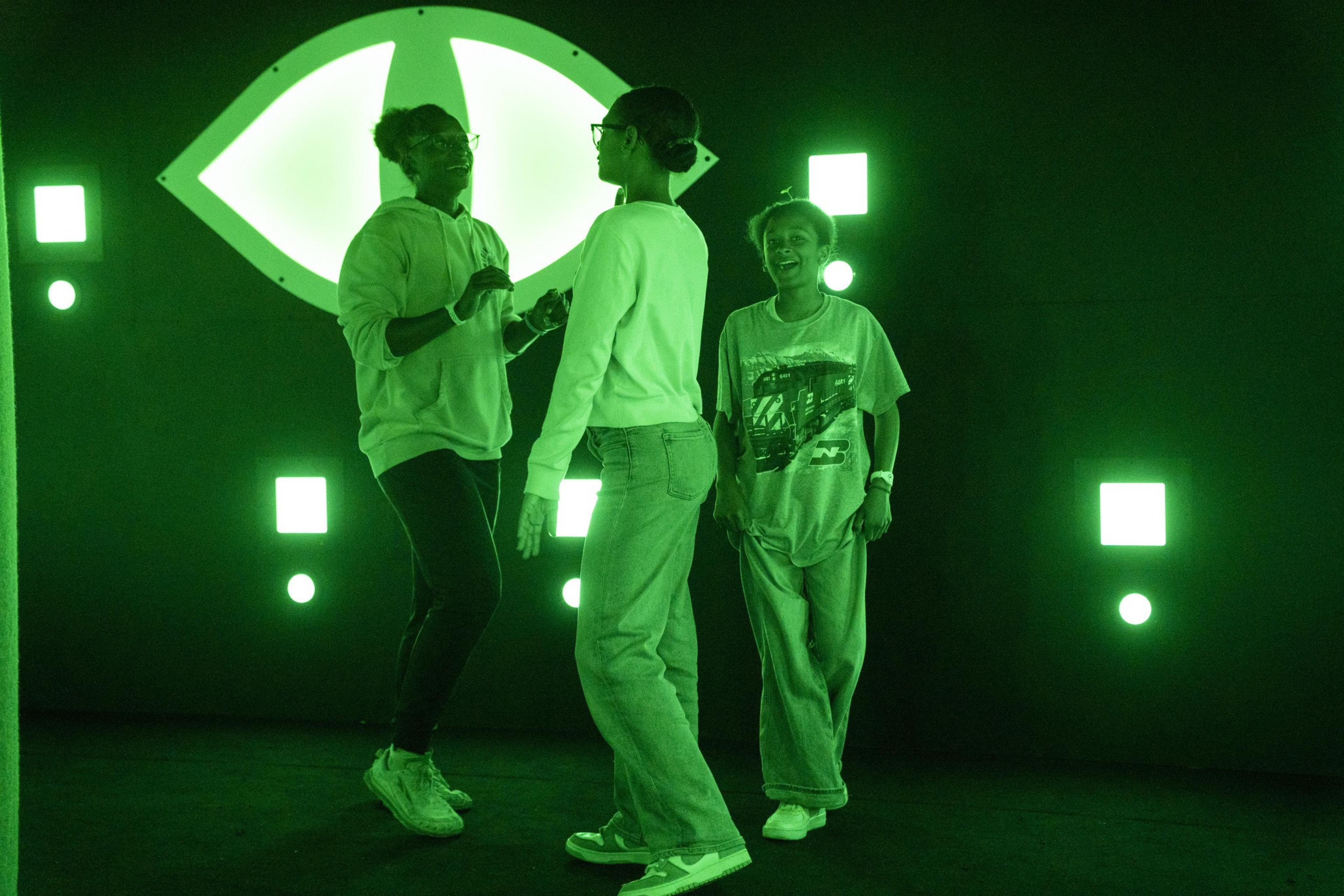 Three people are smiling and interacting in a dark room illuminated by green neon lights and geometric shapes on the walls.