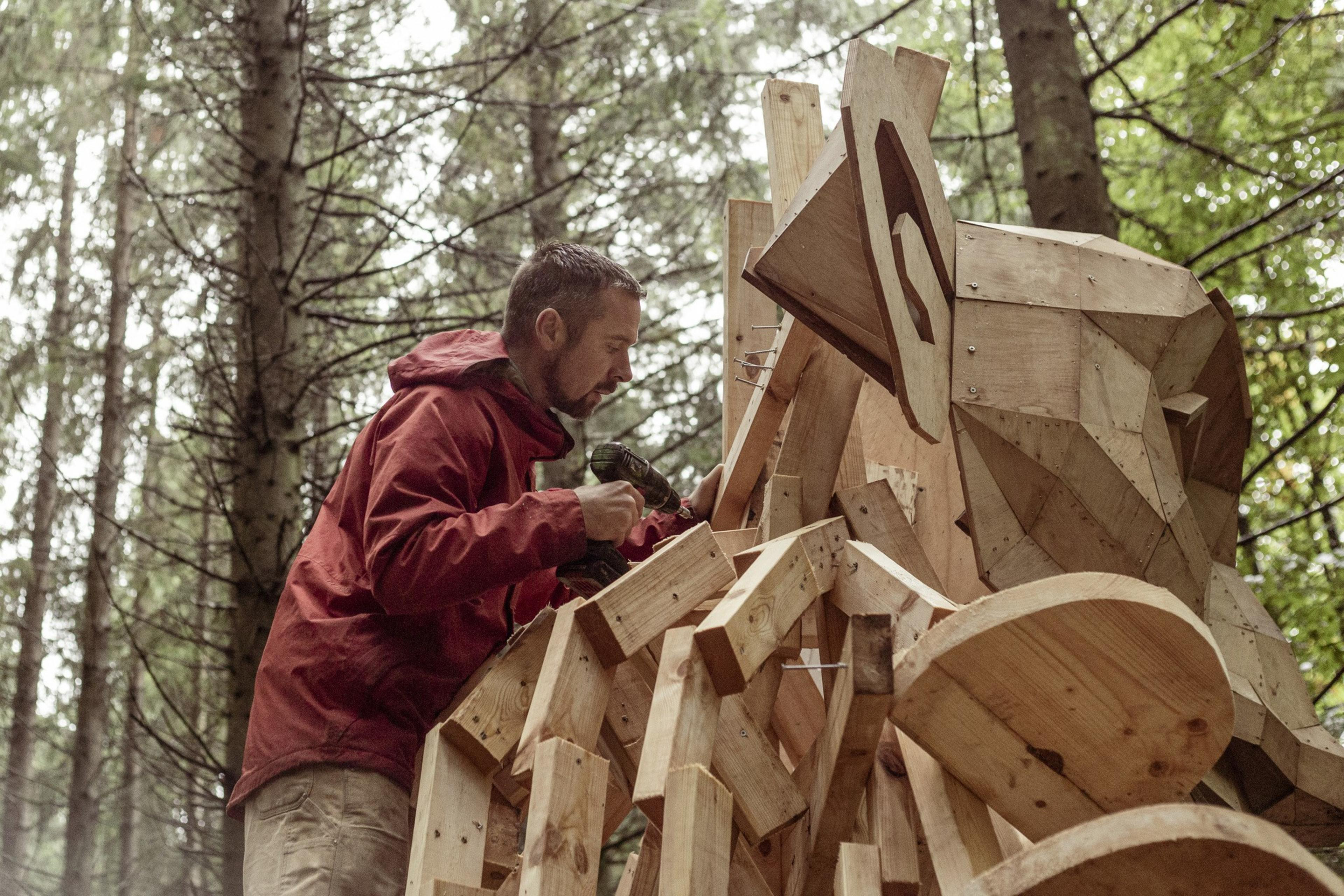 A person in a red jacket is using a power drill to work on a large, wooden sculpture in a forest. The sculpture has geometric shapes and resembles an animal.