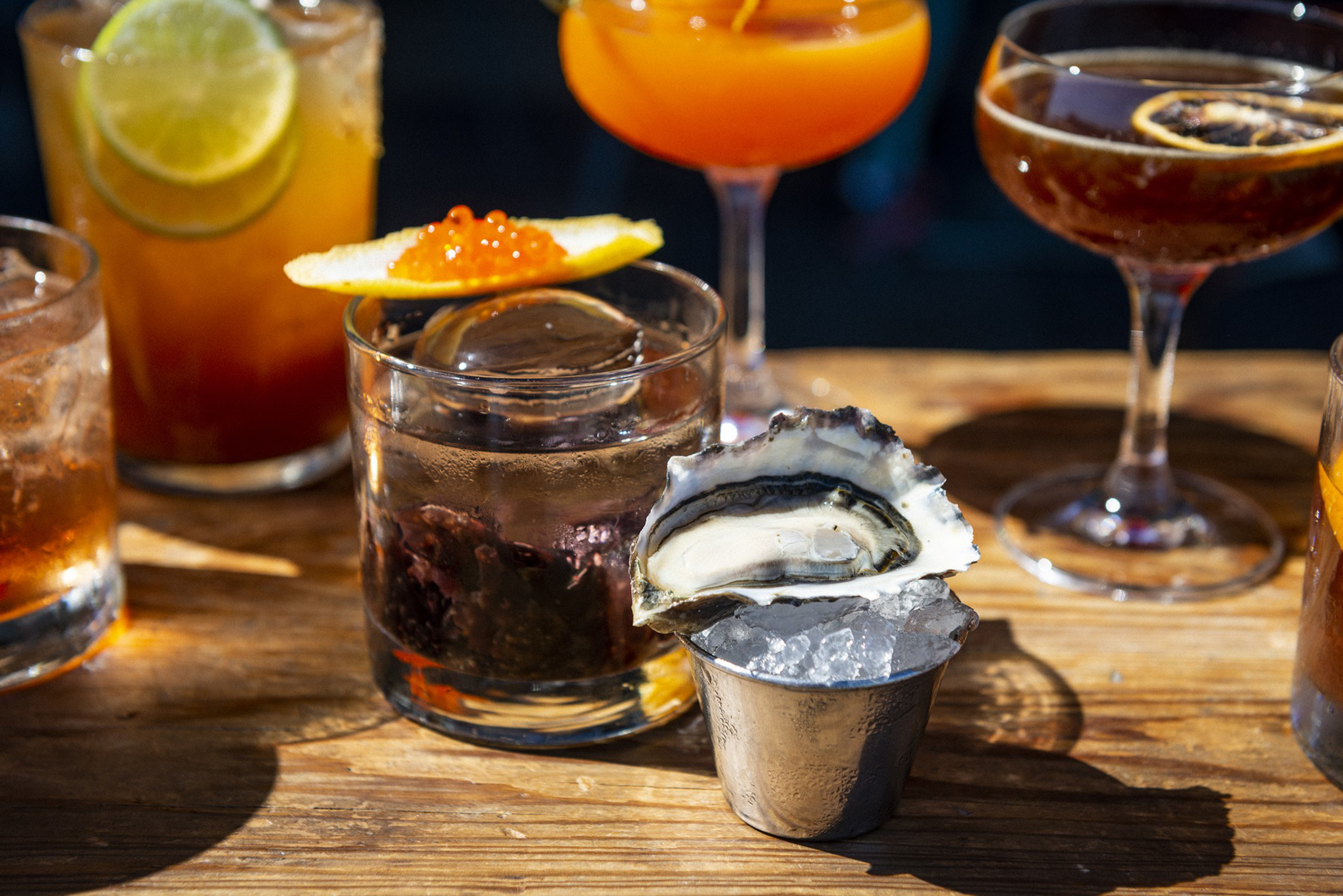 A wooden table holds various cocktails including one with a lemon slice topped with orange roe and a metal cup with an oyster on ice.