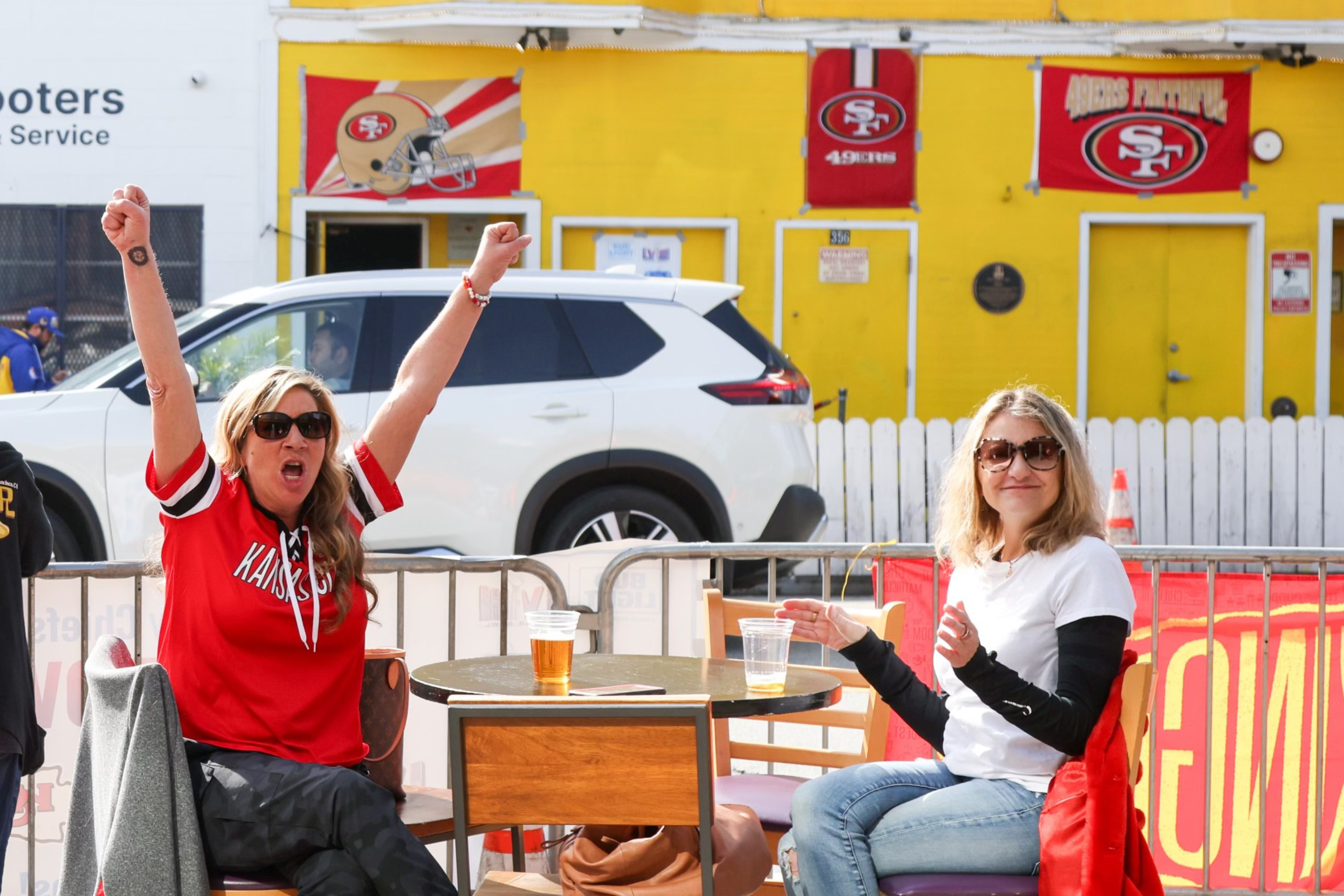 Two women at an outdoor table, one cheering with raised arms; a yellow building with sports banners behind them.