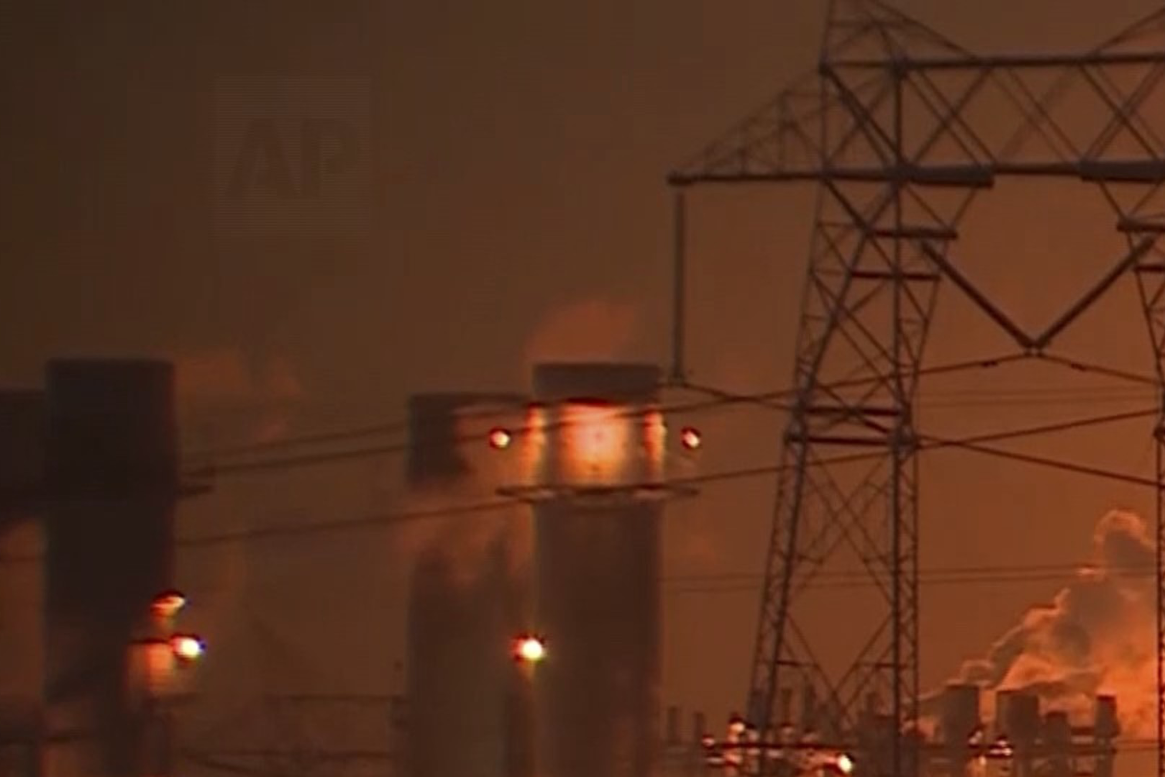 A large industrial complex is engulfed in flames, with bright orange and yellow fire illuminating the night. Power lines and structures are silhouetted.