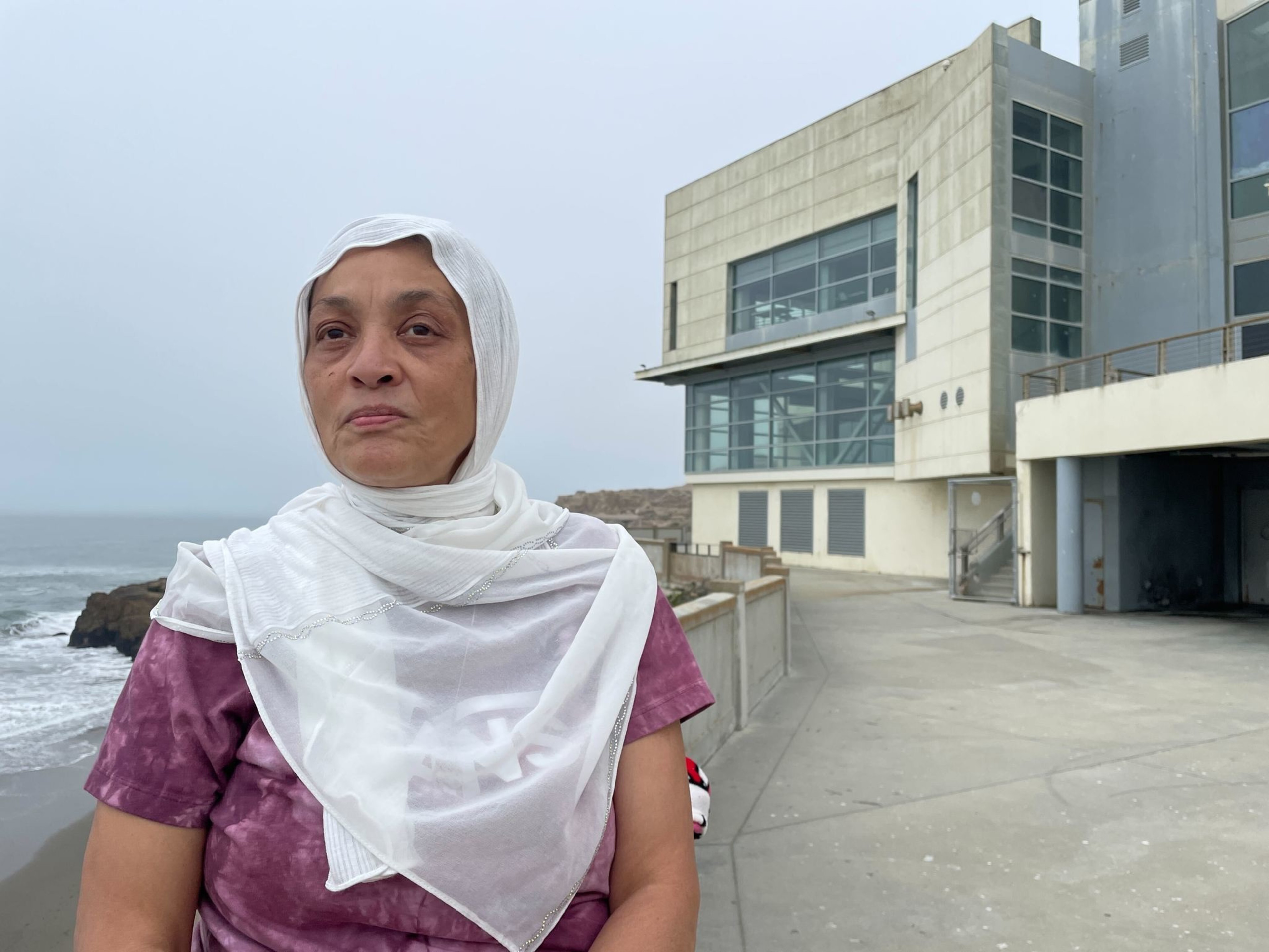 A woman wearing a white headscarf and purple shirt stands near a modern building by the ocean, with cloudy skies and rocky shoreline visible.