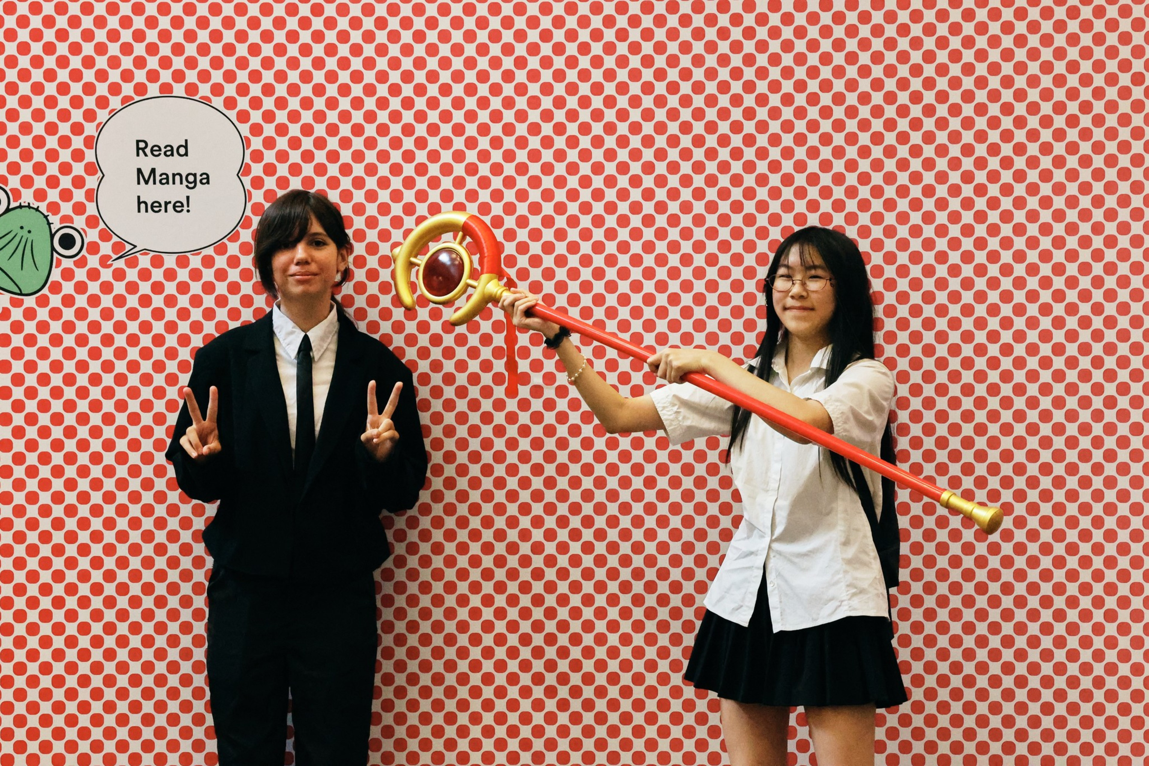 Two people stand in front of a red and white polka dot wall with a speech bubble saying “Read Manga here!”; one holds a staff while the other shows peace signs.