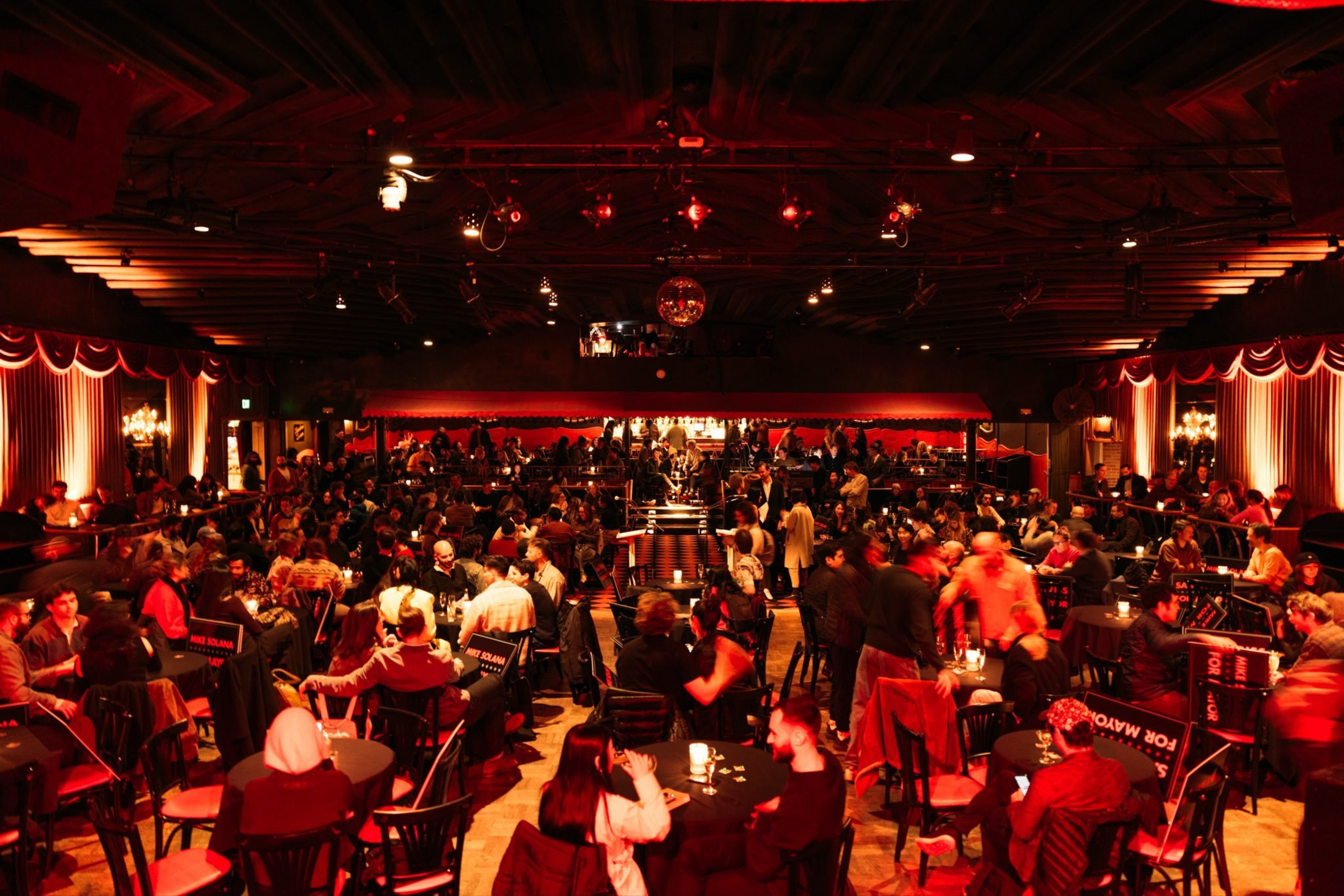A crowded, warmly lit nightclub with round tables, chairs, people socializing, drinks, and red curtains along the walls under wooden ceiling beams.