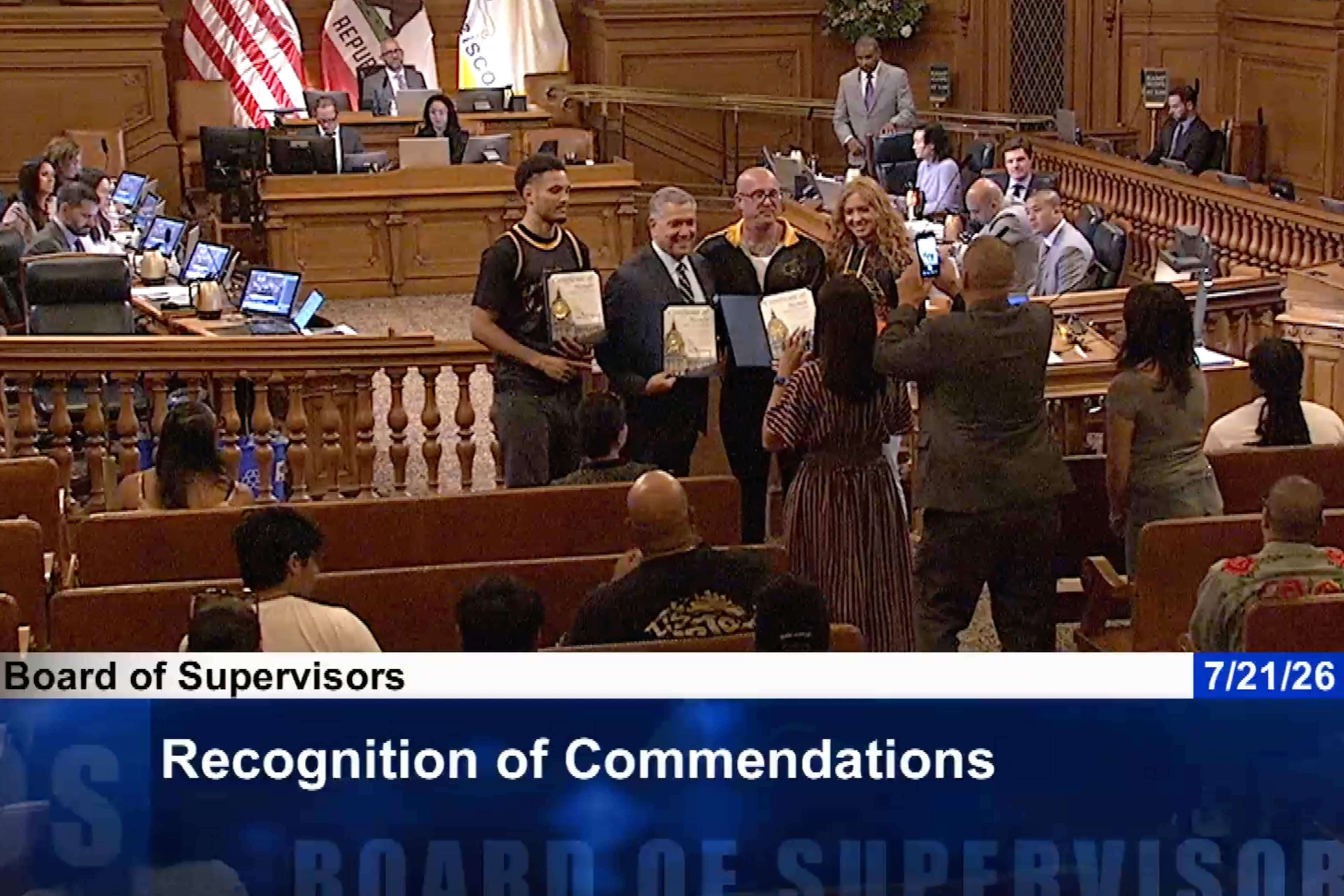 Four people stand holding plaques while others take photos during a Board of Supervisors meeting, with officials seated at desks in the background.