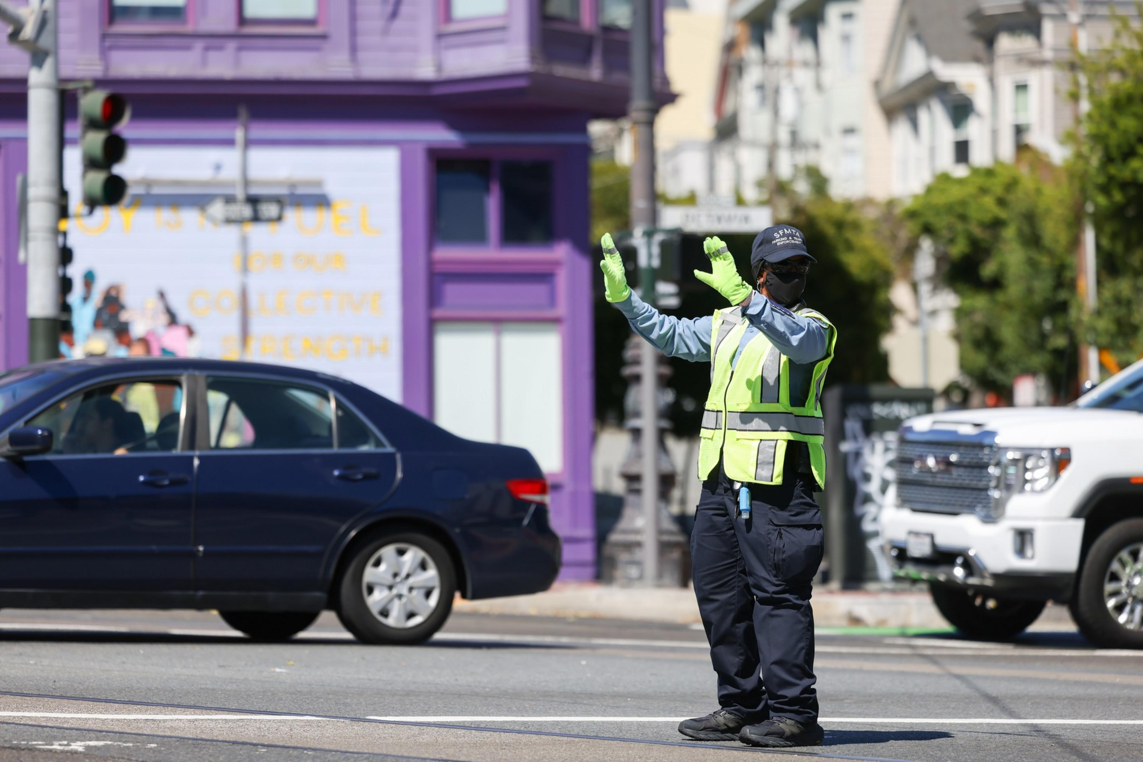 A traffic officer wearing a mask and neon gloves directs cars on a street with a purple building and a white pickup in the background.