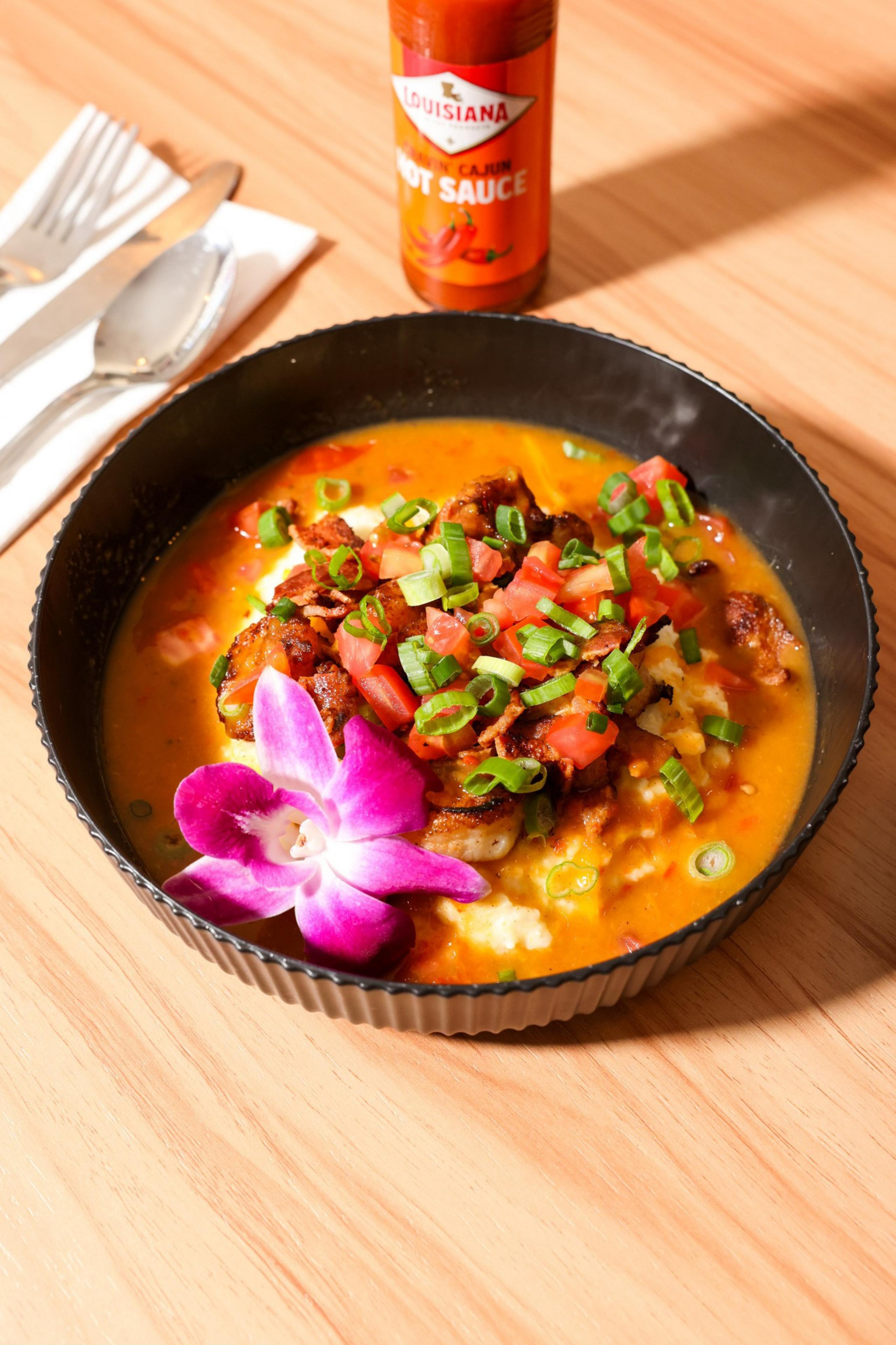 A black bowl holds a vibrant dish with creamy grits, topped with diced tomatoes, green onions, and fried bits, garnished with a purple orchid and hot sauce nearby.