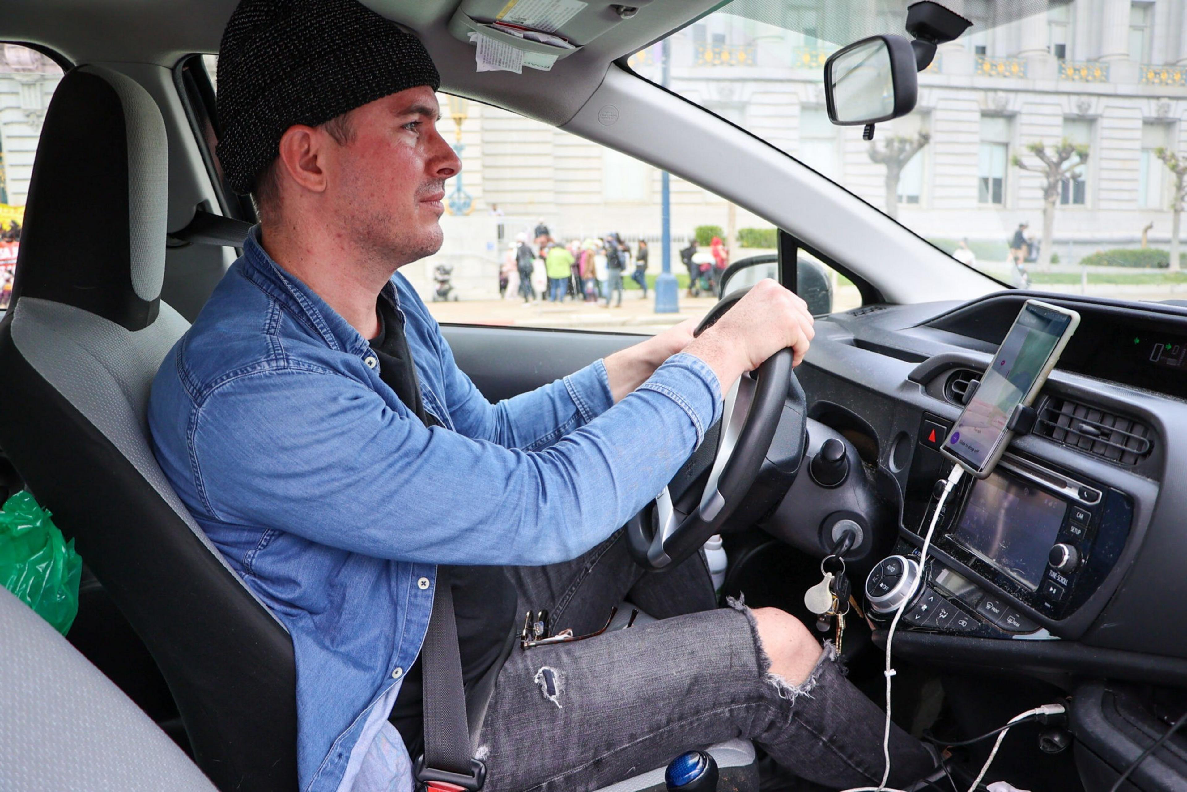 A man wearing a black beanie, blue denim jacket, and ripped jeans is sitting in the driver's seat of a car, holding the steering wheel with both hands.