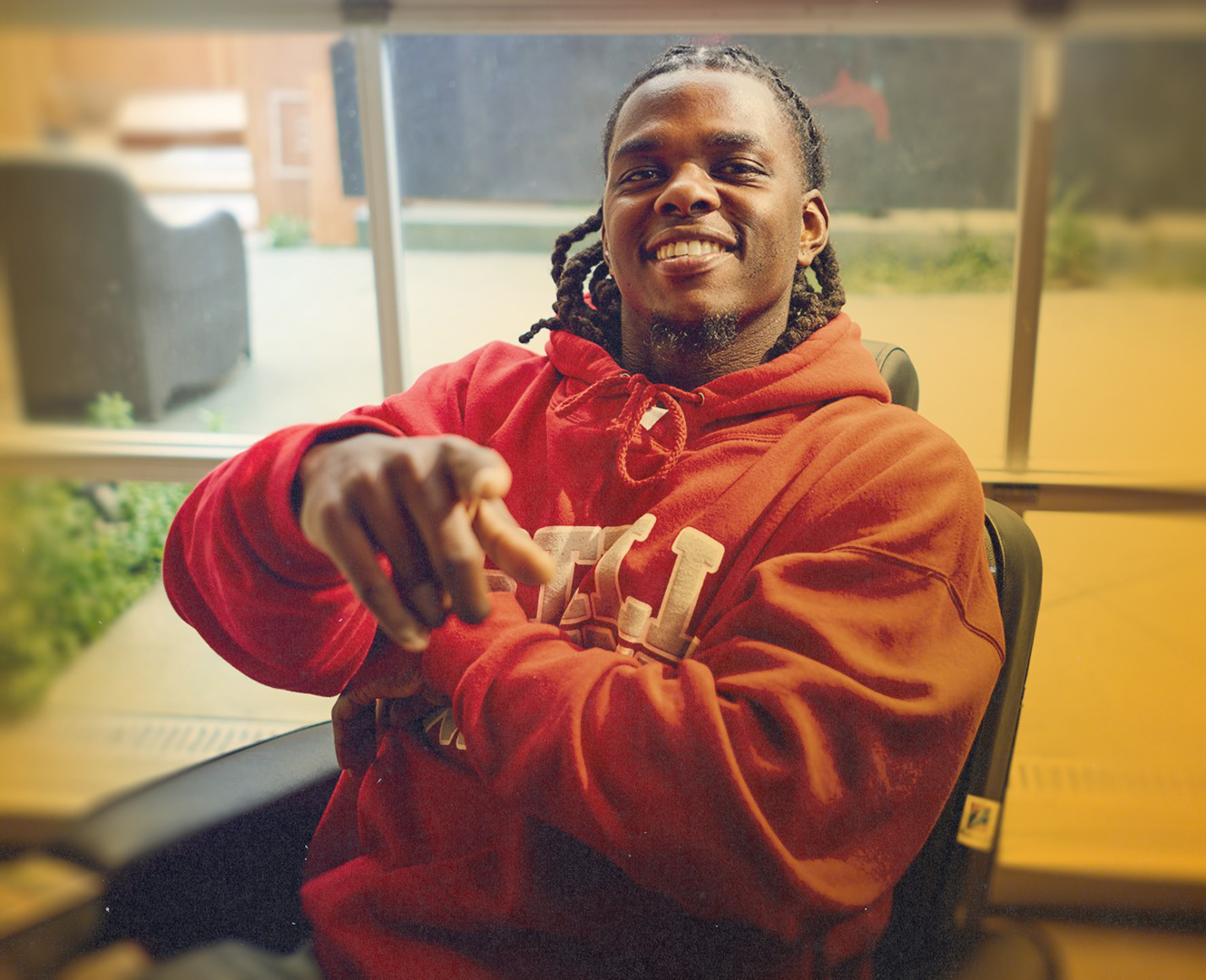 A smiling man with braided hair wears a red hoodie and points forward while sitting in a chair in a warmly lit indoor space.