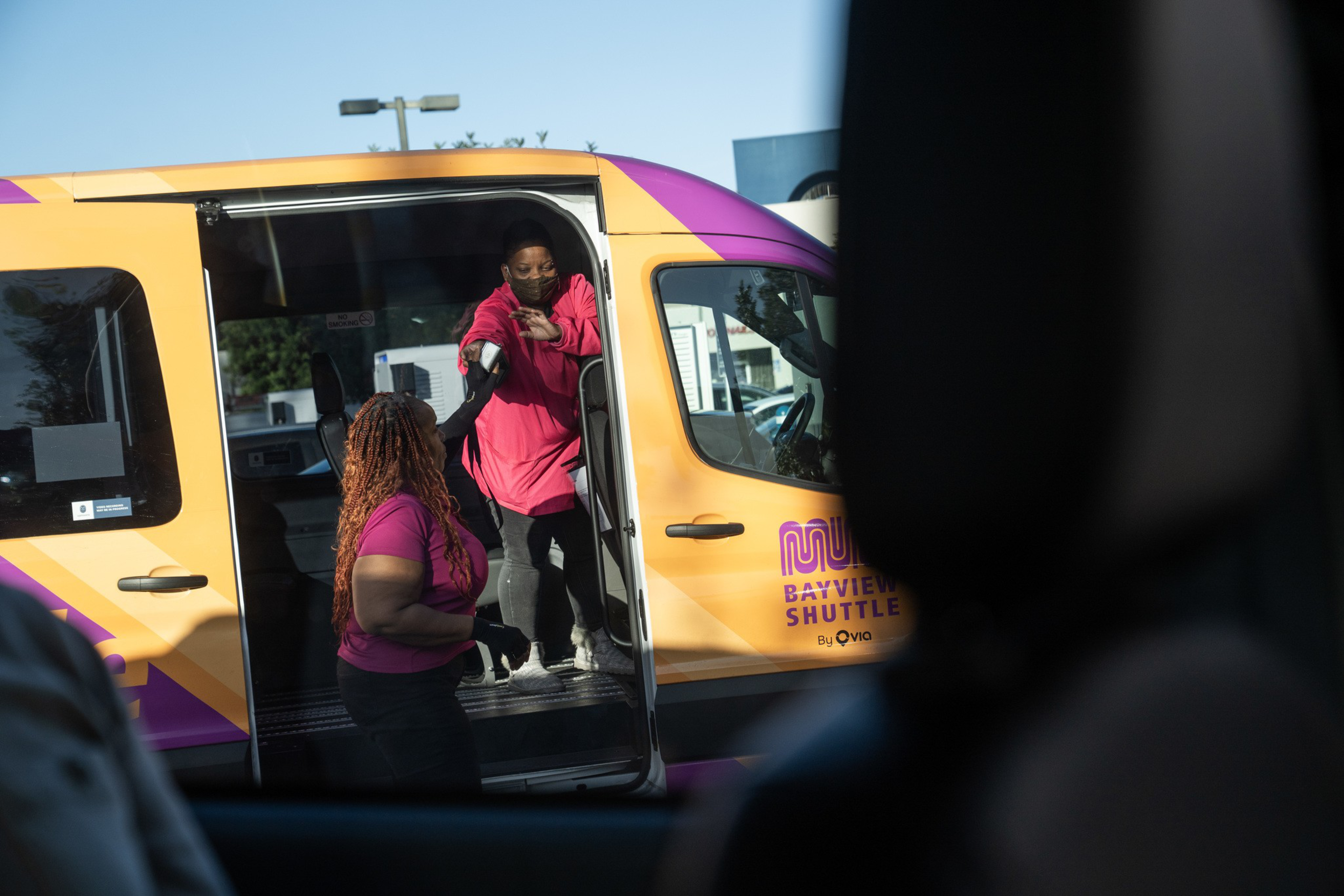 A woman in a pink top reaches out from a yellow and purple shuttle van toward another woman standing outside the van.