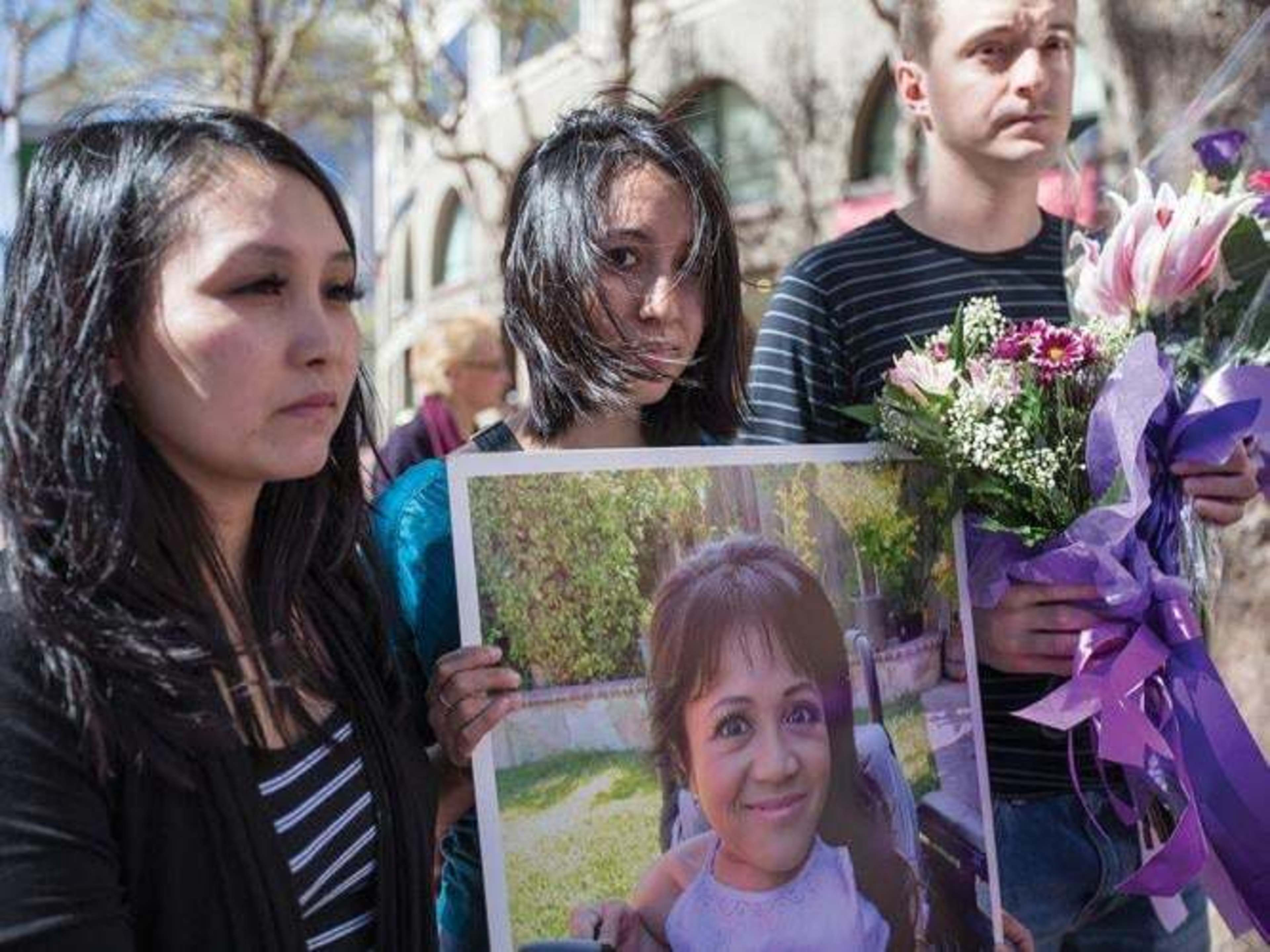 Three somber individuals hold a person’s photo and flowers with purple ribbons.