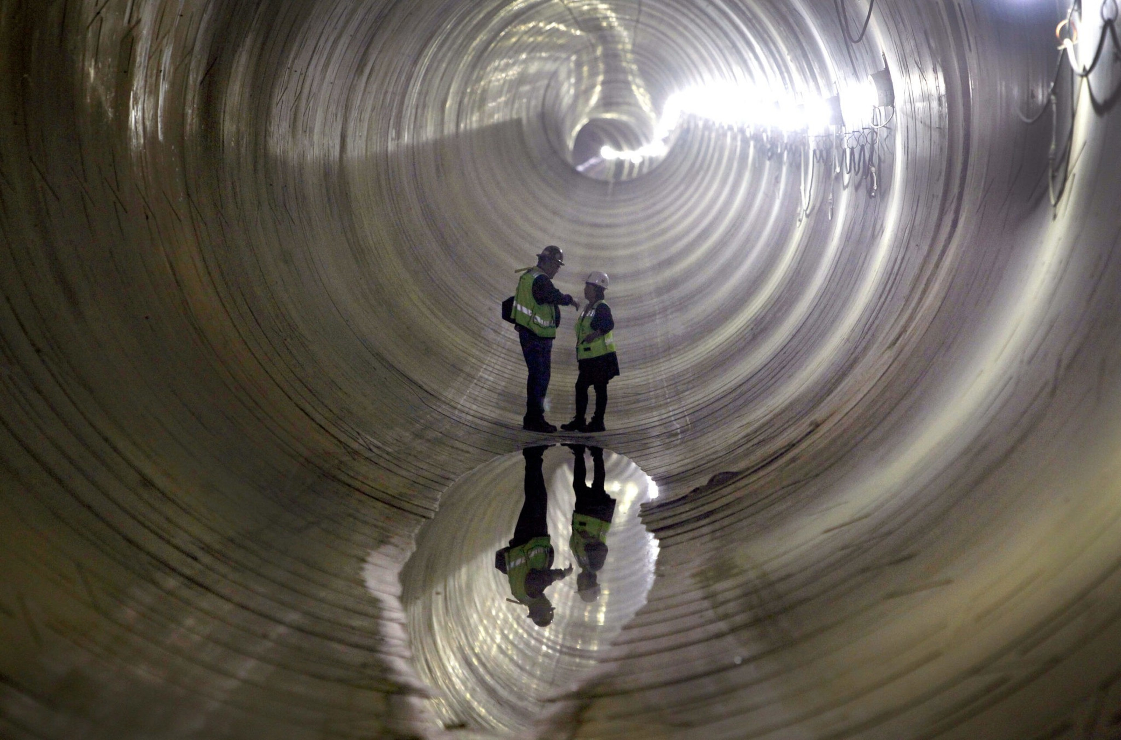 Two workers wearing helmets and reflective vests stand inside a large, cylindrical tunnel with light reflecting off a puddle below them.