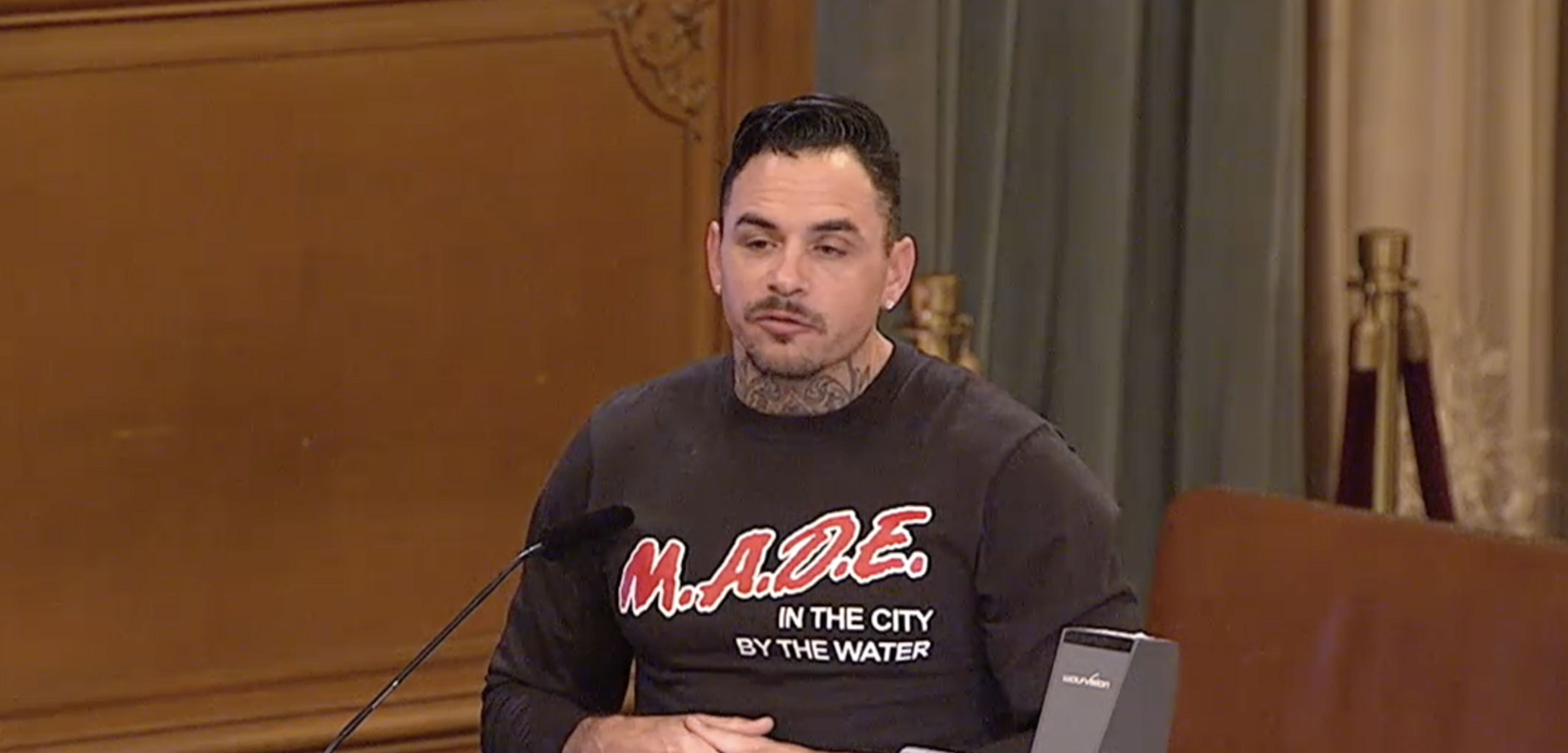 A man stands speaking at a podium, wearing a black shirt with red and white text "M.A.D.E. in the City by the Water" in a wood-paneled room with curtains.