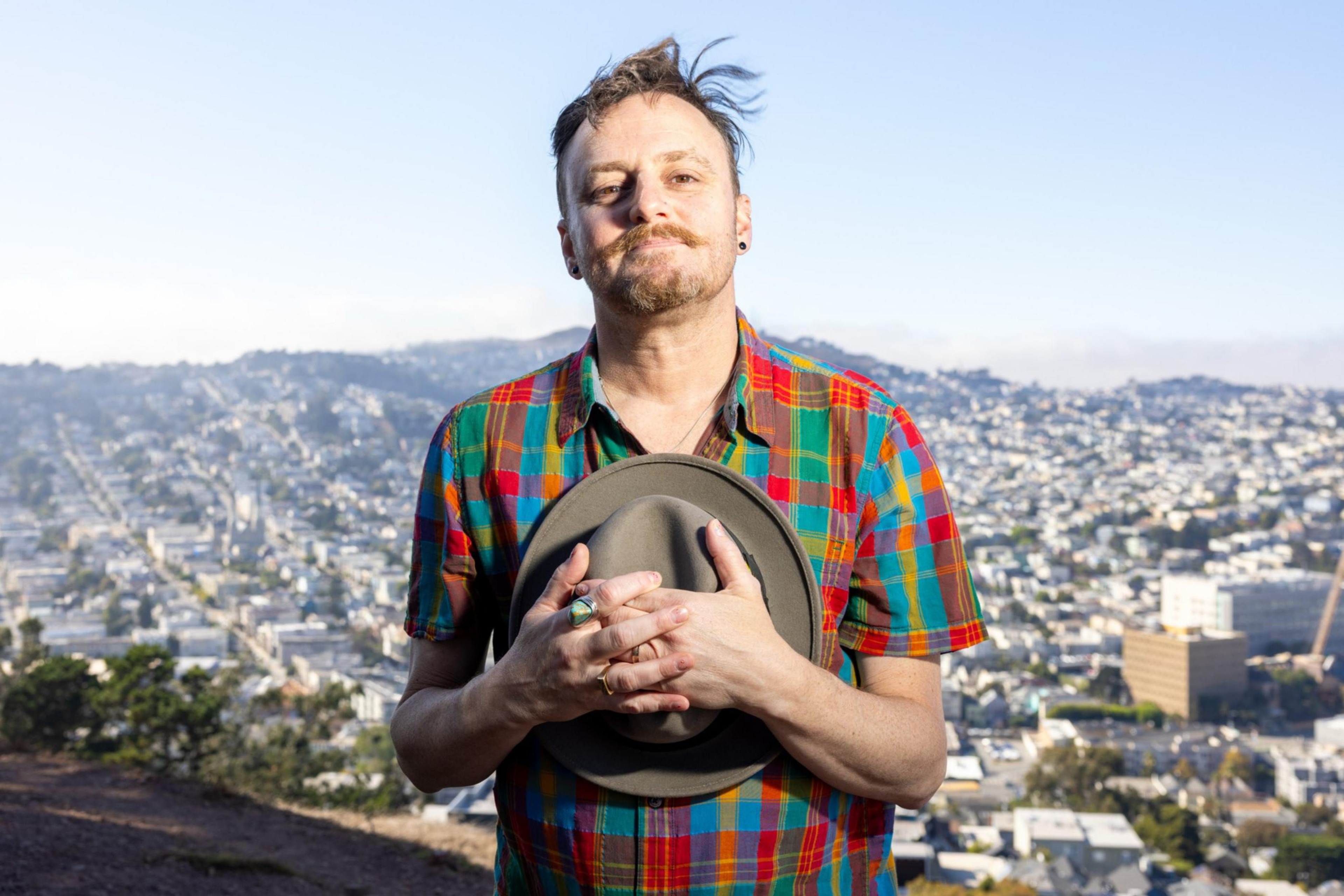A man with a mustache and short hair holds a hat to his chest, wearing a colorful plaid shirt, standing outdoors with a cityscape and hills in the background.